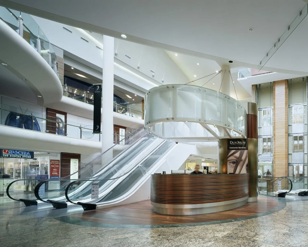 Interior of a modern shopping mall featuring an escalator, a curved reception or information desk, and multiple levels with storefronts and glass railings.