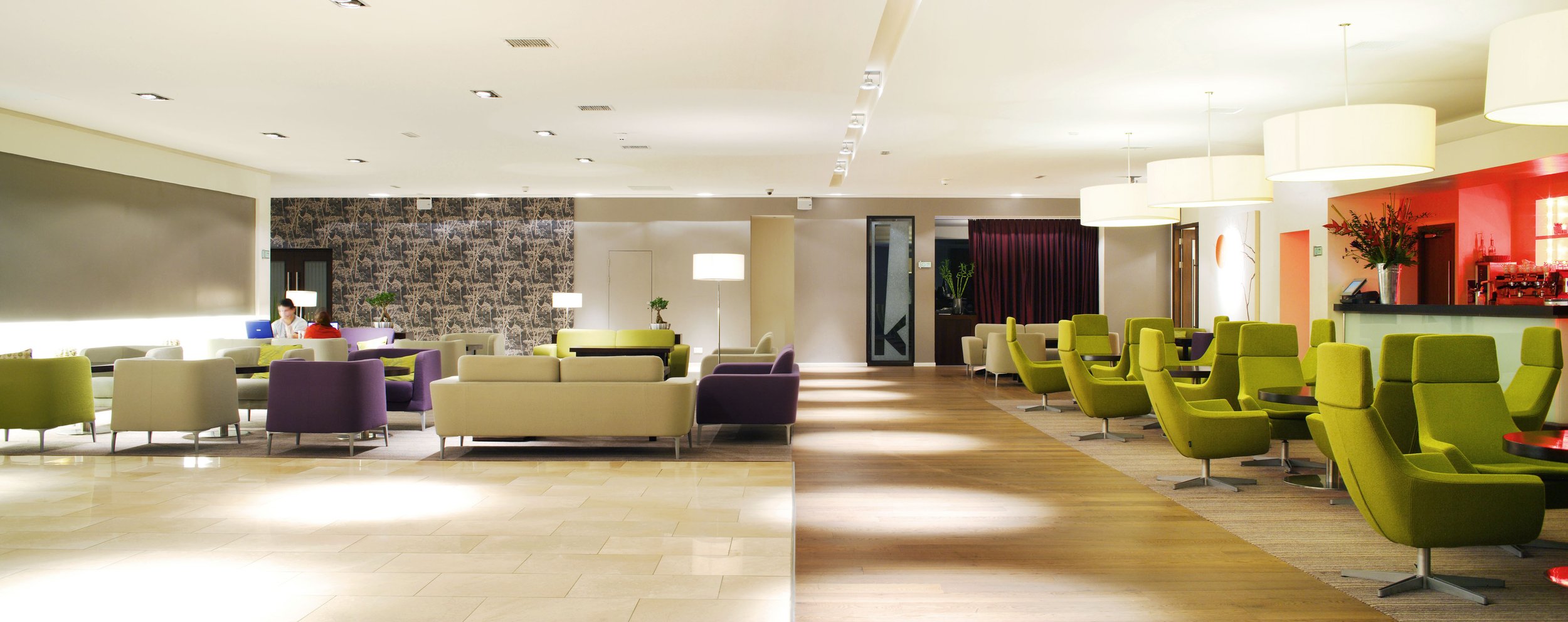Modern hotel lobby with colorful armchairs and sofas, large white pendant lights, wood and tile flooring, and a bar with red lighting and floral arrangements.