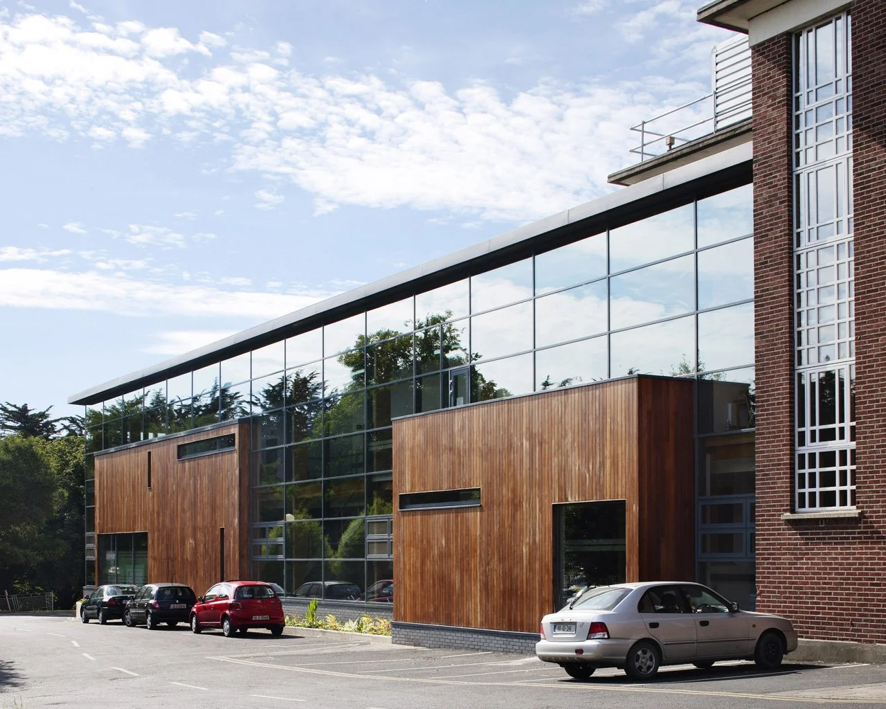 Modern building with large glass windows and wooden paneling in a parking lot with cars, under a partly cloudy sky.