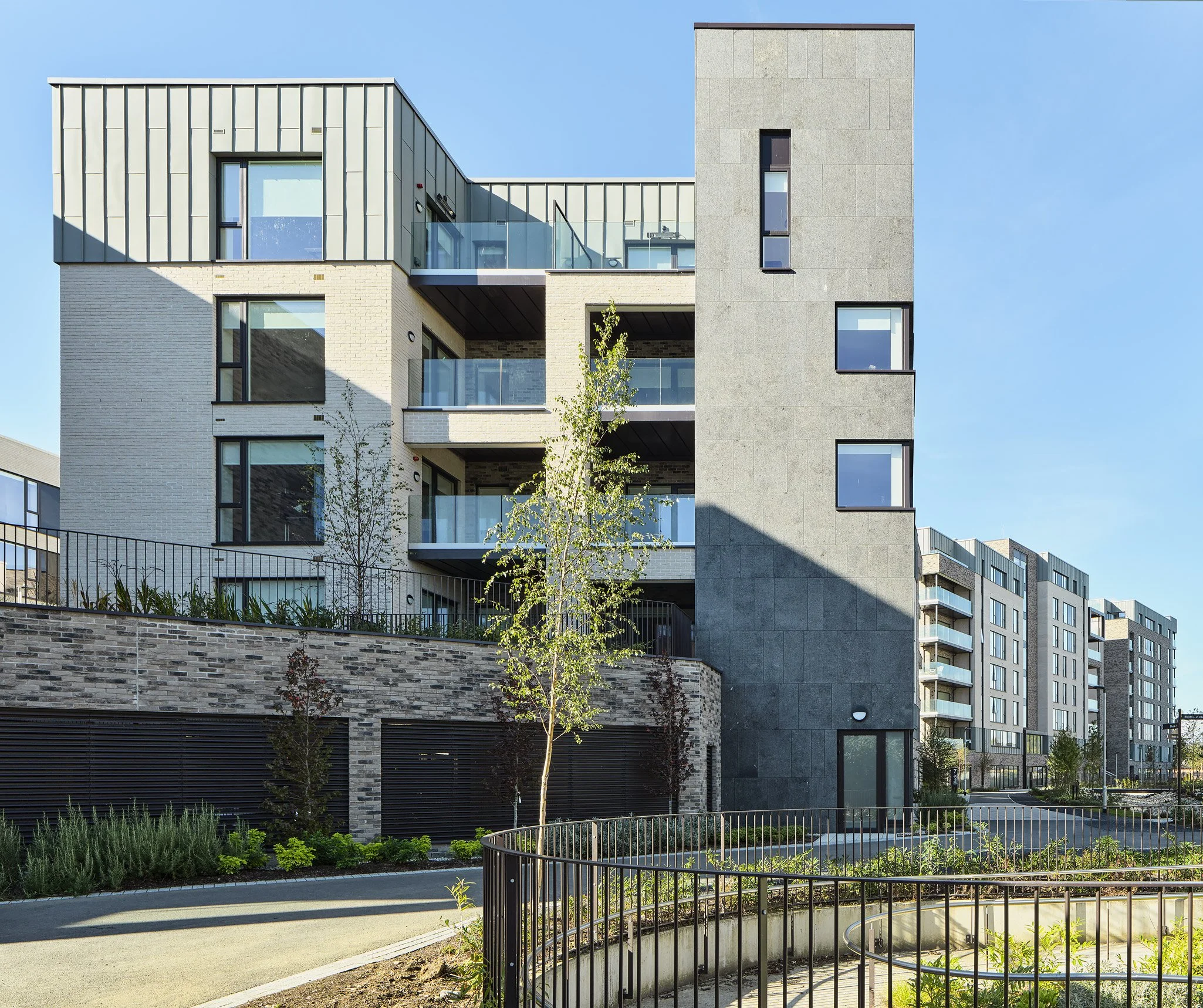 Modern multi-story apartment building with balconies and large windows, landscaped outdoor area, and clear blue sky.