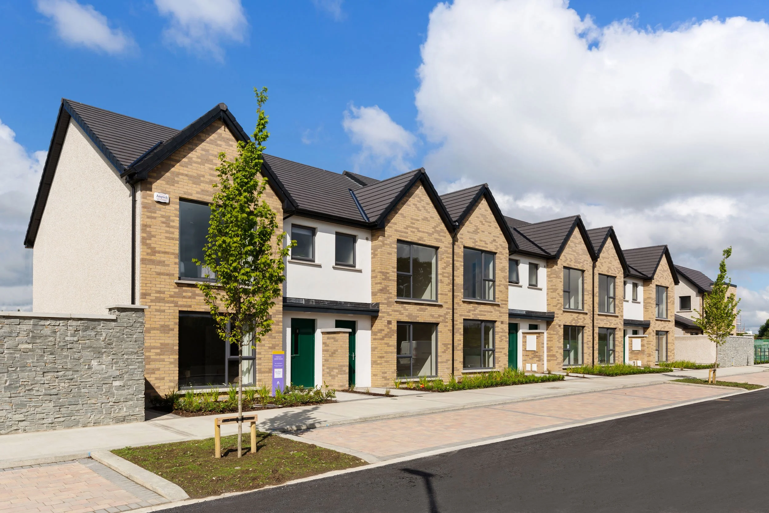 Modern row of townhouses with brick and white walls, large windows, small front gardens, and young trees under a blue sky with clouds.