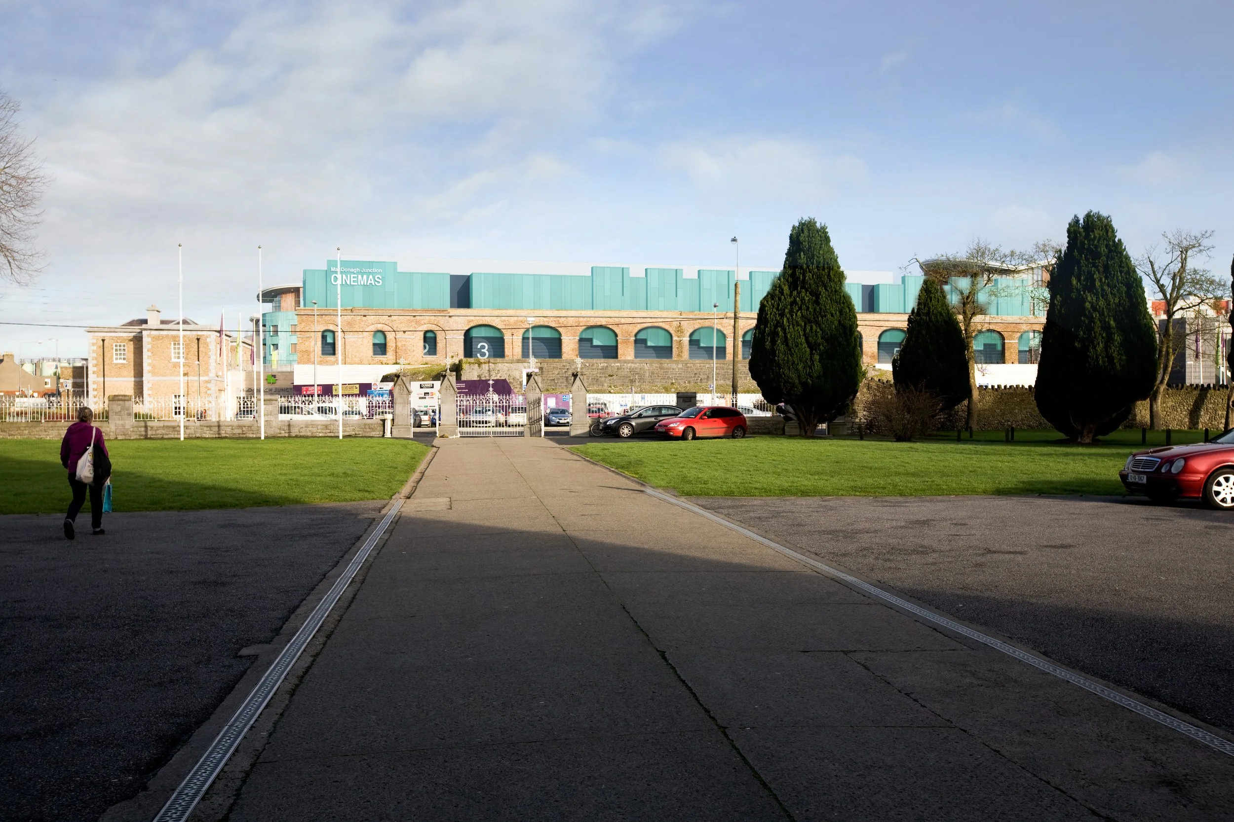 View of a park with a paved walkway, green grass, trees, and parked cars, with a building labeled "CINEMAS" in the background under a partly cloudy sky.