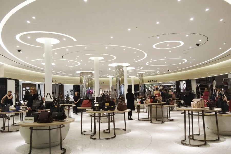 Inside a luxury designer handbag store with circular display tables showcasing handbags, mannequins dressed in designer clothing, and illuminated store signage for Prada and Tom Ford, with several customers browsing.