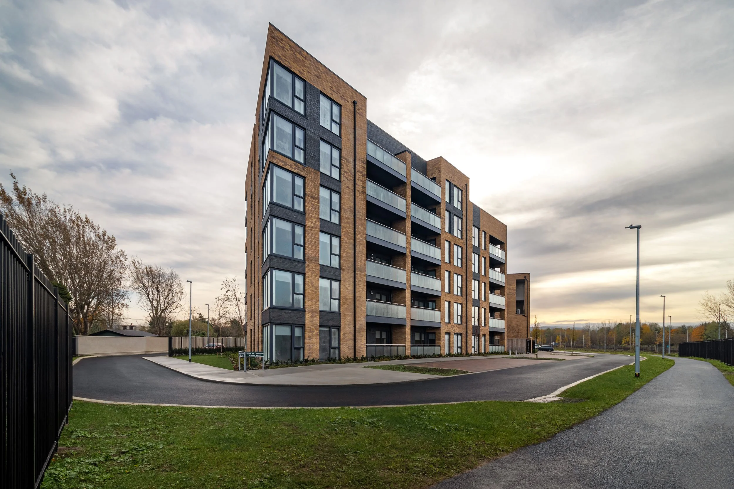 A modern multi-story apartment building with brick and black exterior, featuring large glass windows and balconies, set in a landscaped area with a curved driveway and pathway, under a cloudy sky.