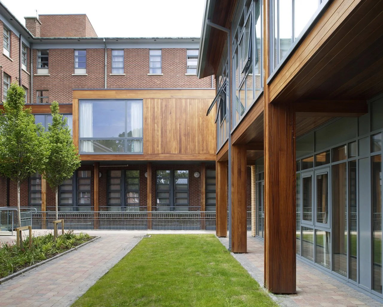 Modern building with brick, glass, and wood exterior, green lawn, paved walkway, and trees.