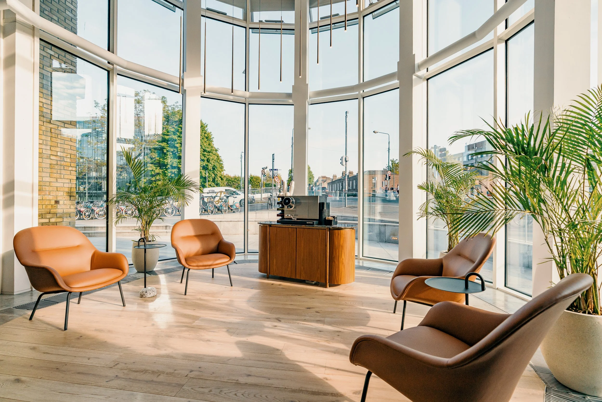 Modern coffee shop interior with large floor-to-ceiling windows, several tan leather chairs, small round tables, potted plants, and a wooden counter with a coffee machine on top.