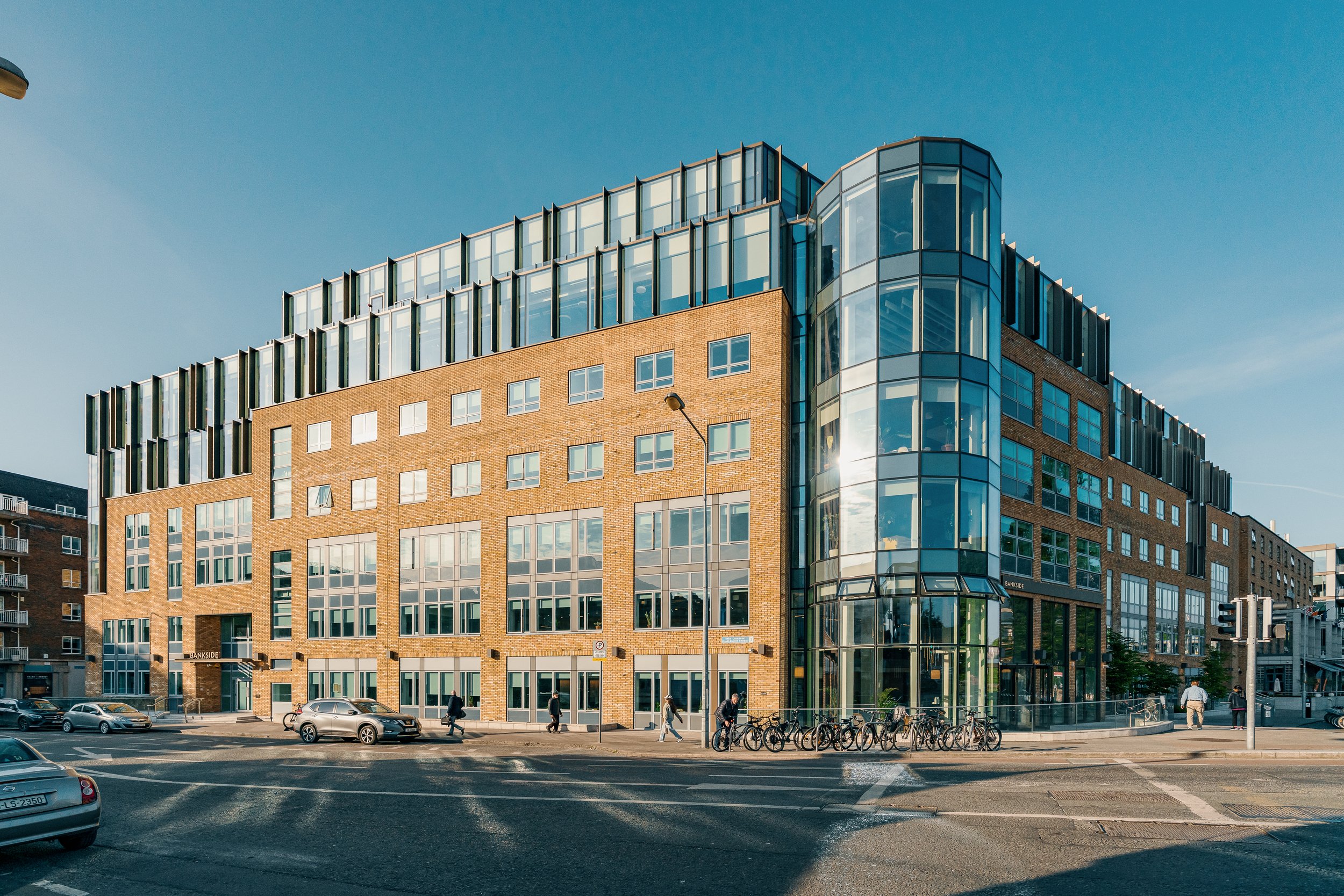 Modern multi-story building with brick and glass exterior, bicycles parked outside, people walking, and cars on the street under a clear blue sky.