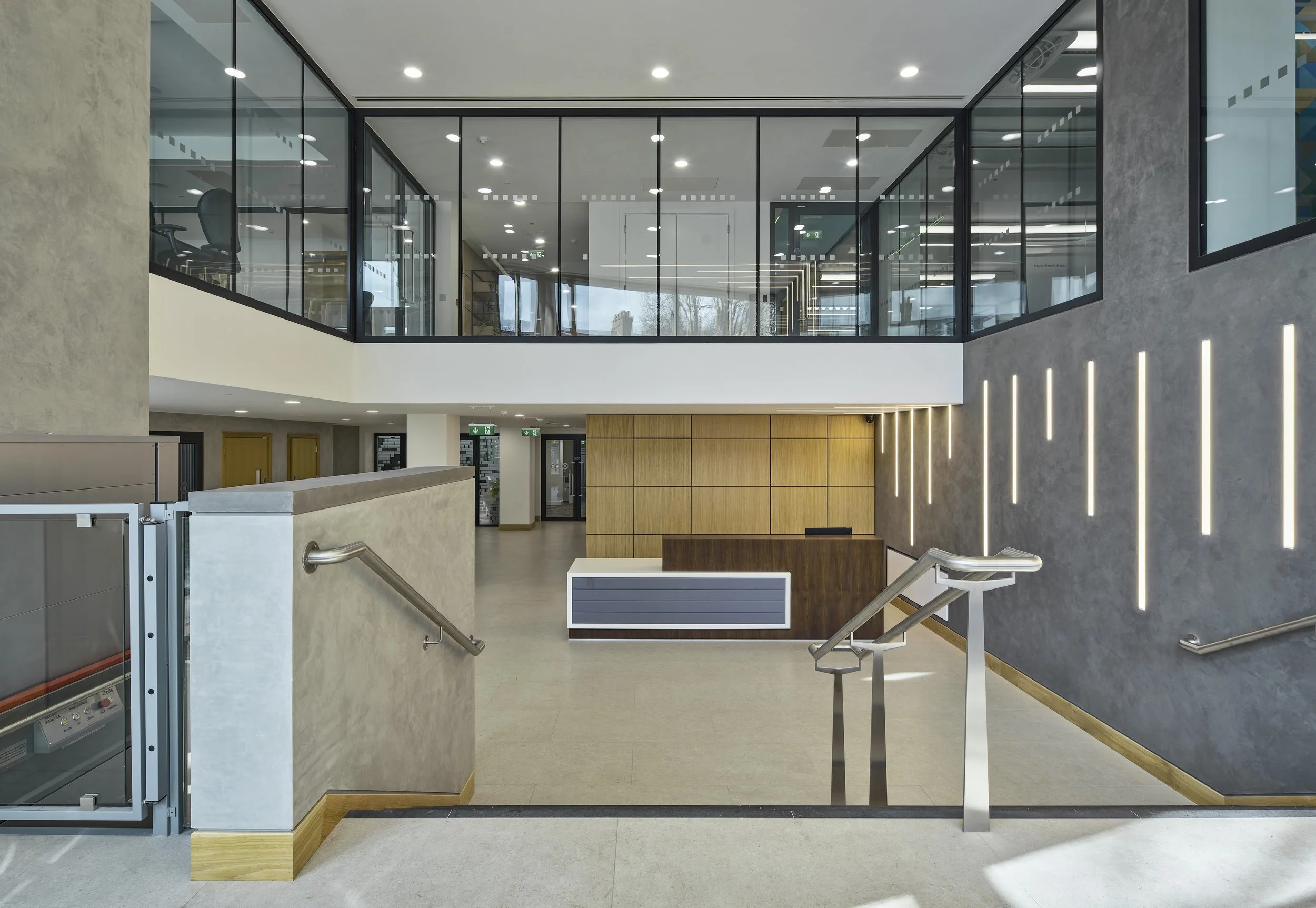 Modern office lobby with wooden reception desk, glass upper floor walls, gray textured walls with vertical light accents, beige flooring, and staircase railing.