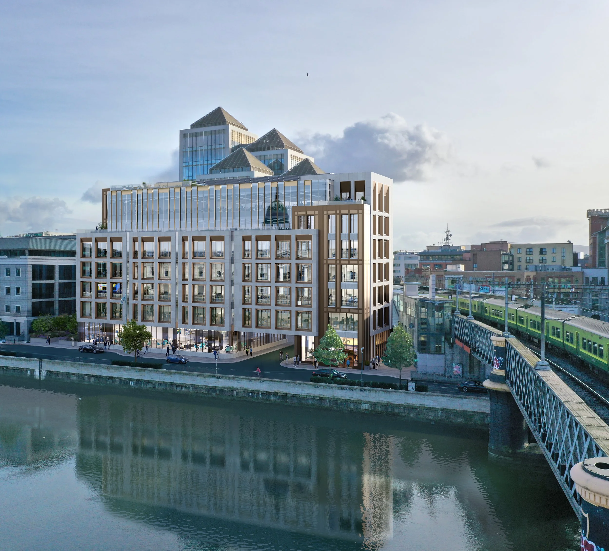 A modern multi-story building situated along a waterway with a bridge and train to the right, and a cityscape with high-rise buildings in the background.