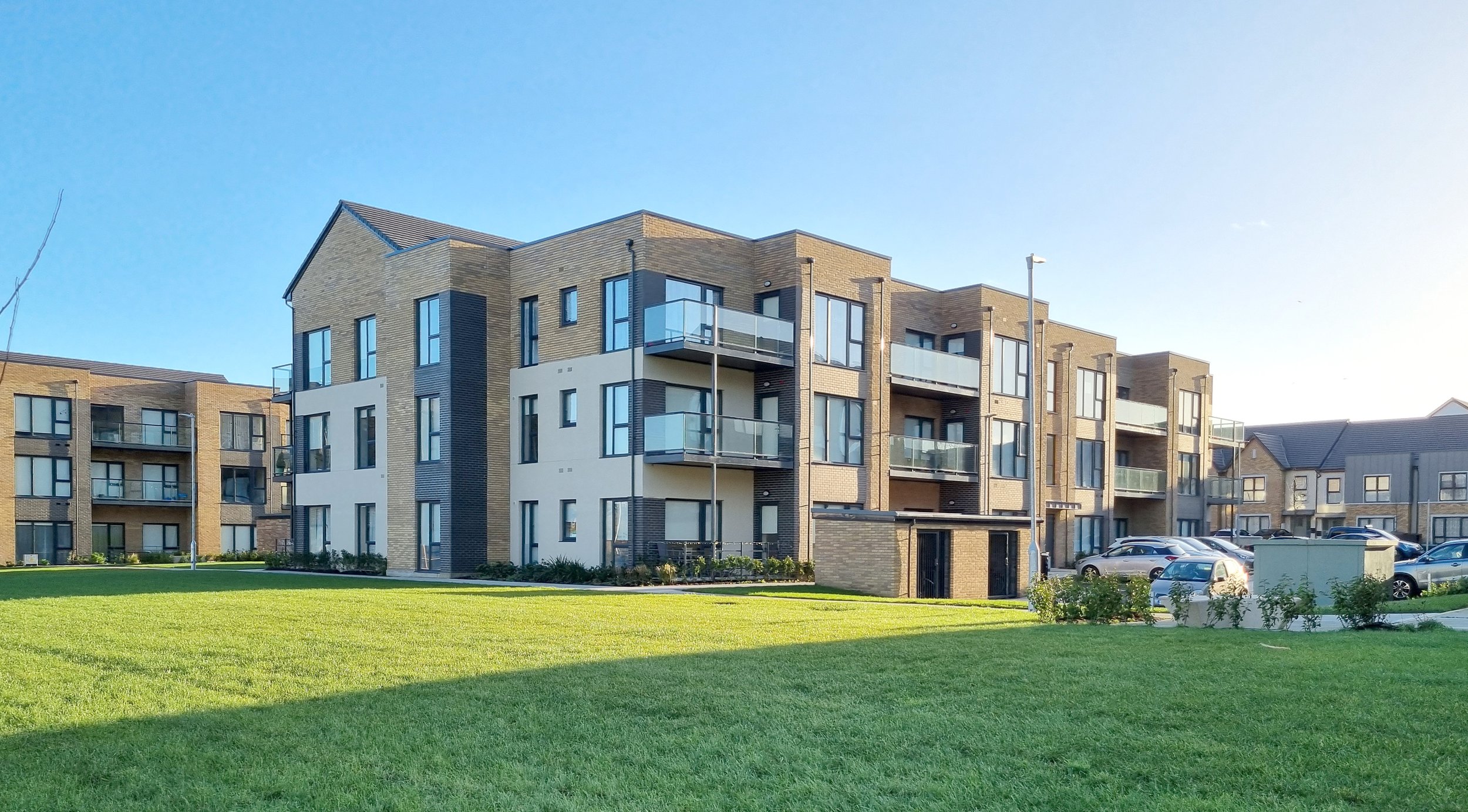 A modern apartment complex with multiple stories, balconies, and parking lot, surrounded by green lawn and clear blue sky.