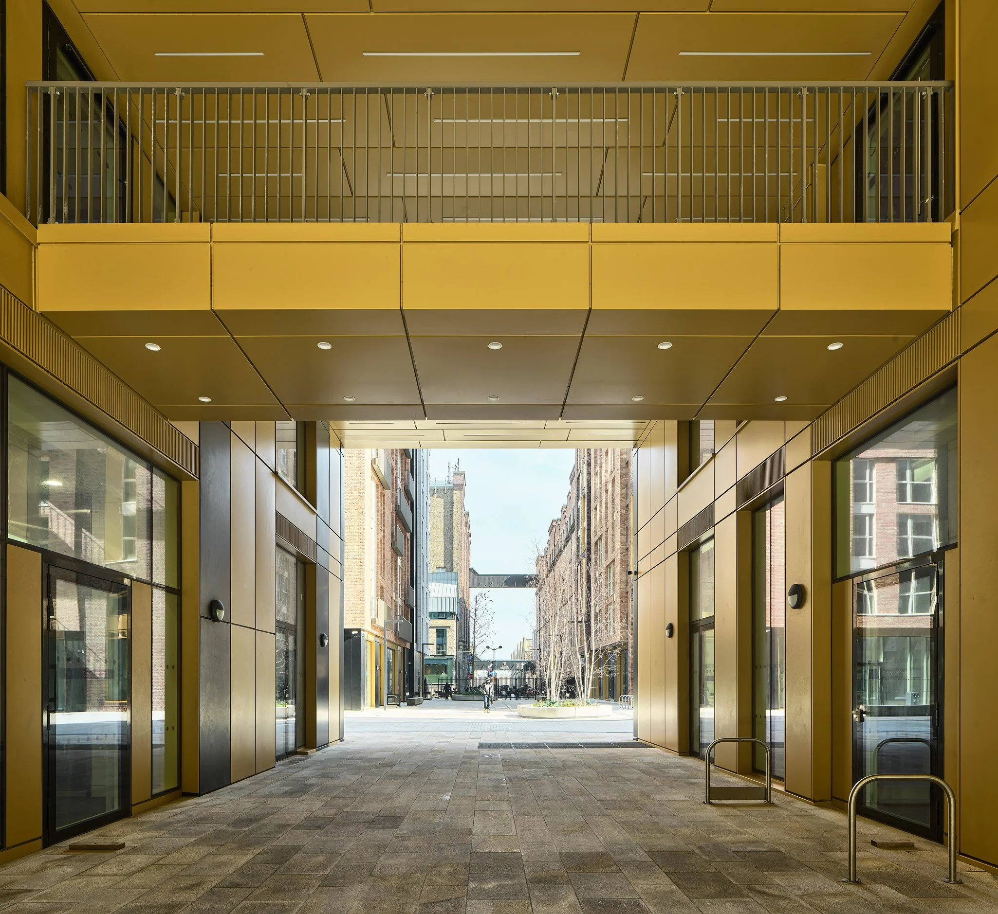 View of a modern urban passageway with yellow and black exterior walls, glass doors, and a balcony above, leading to a city street with buildings and pedestrians in the background.