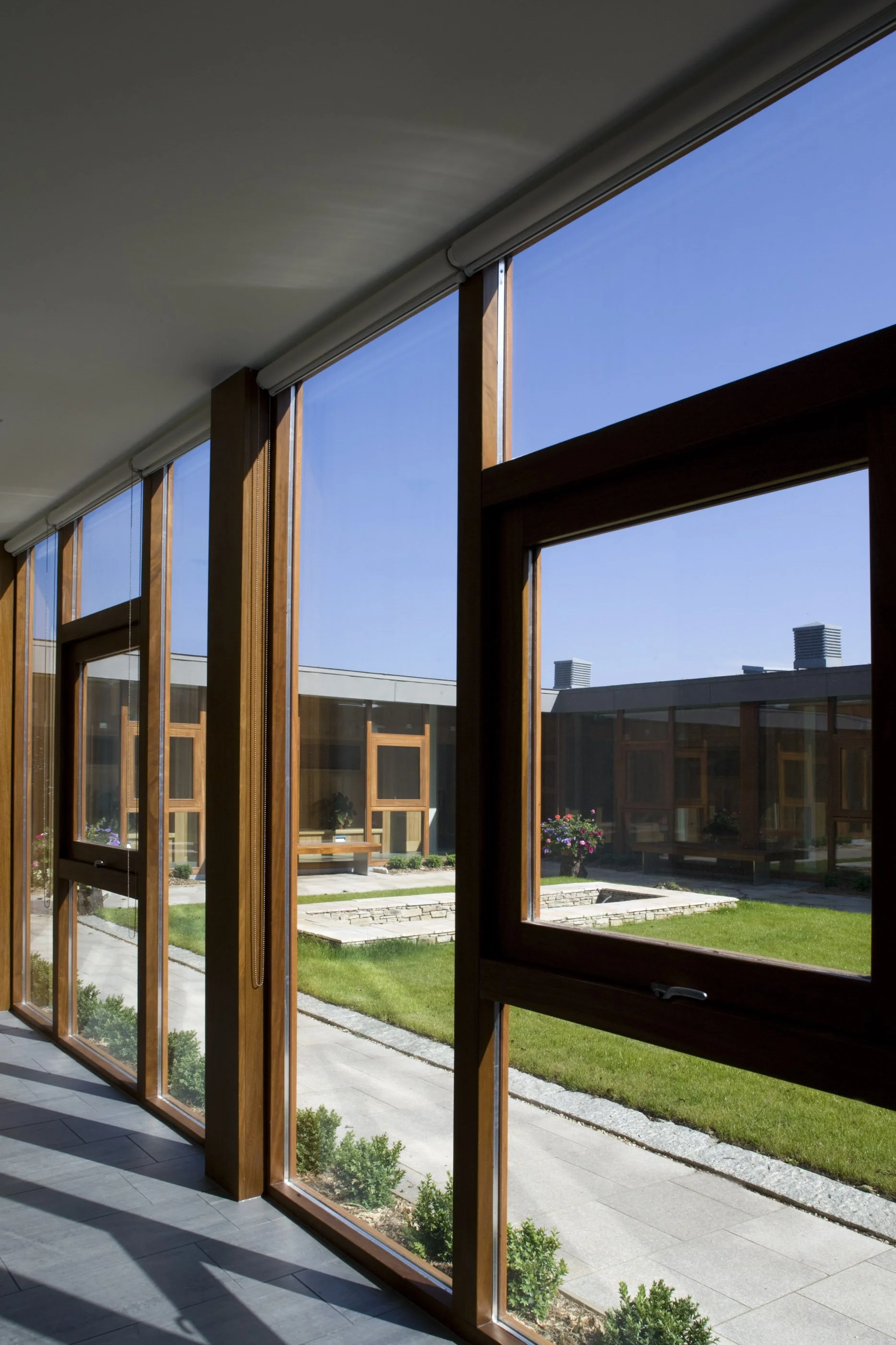 View through large wooden-framed windows overlooking a courtyard with grass, flower pots, and a walkway, under clear blue sky.