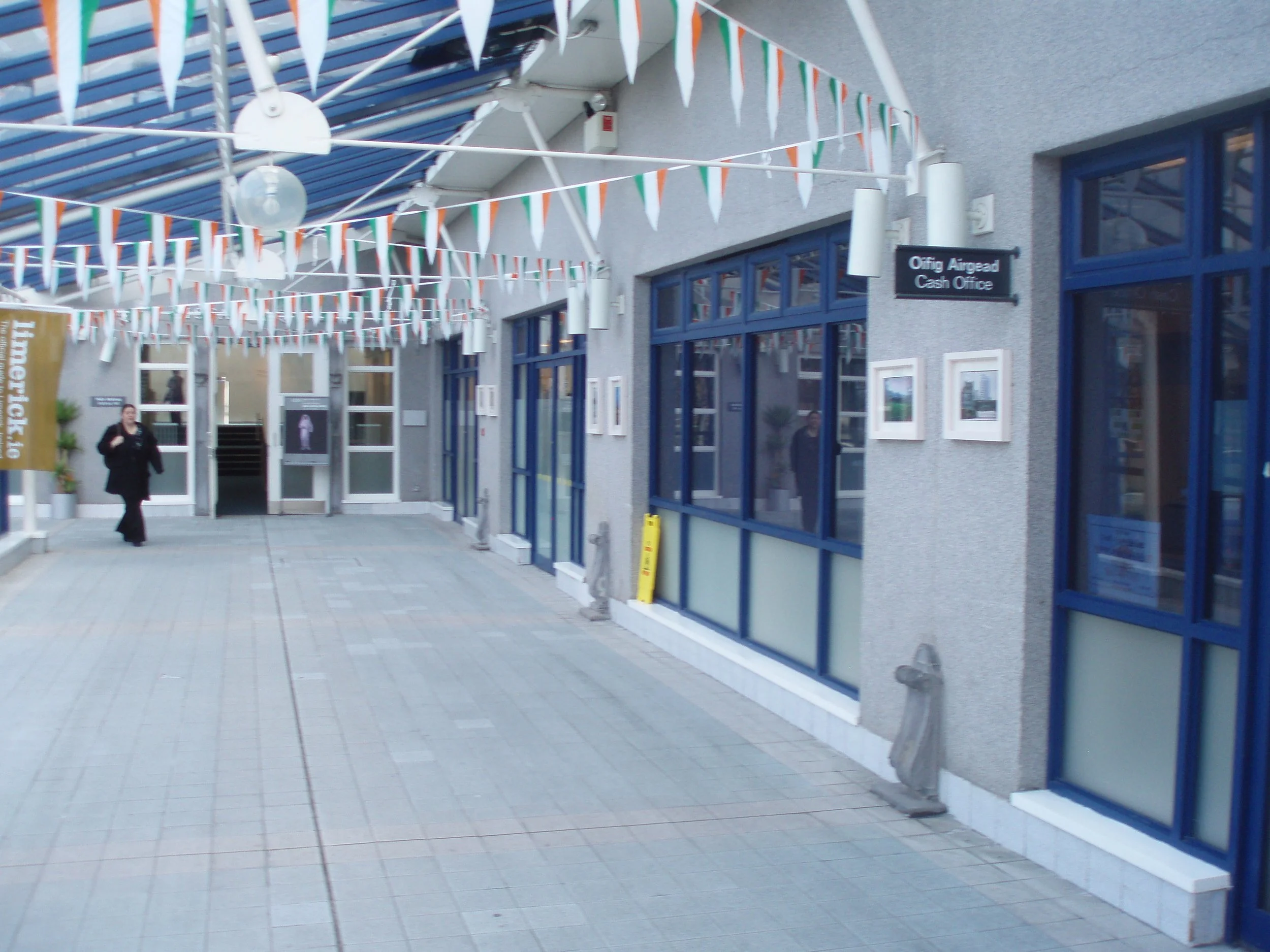 Outdoor area with flags and banners, a woman walking, and a building with blue-framed windows labeled 'Offing Airgead Cash Office'