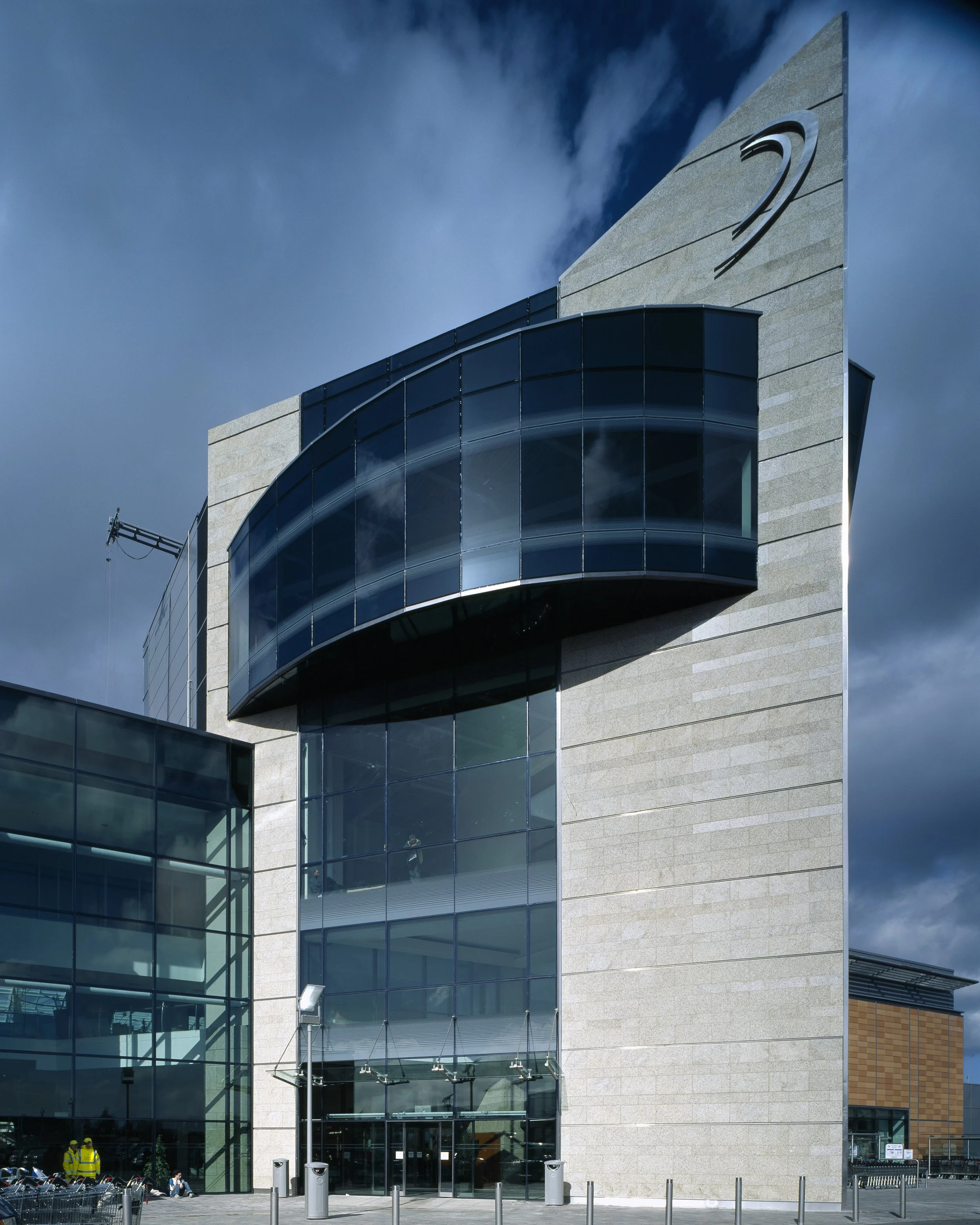 Modern building with glass facade and a logo resembling a stylized swoosh, under a cloudy sky.