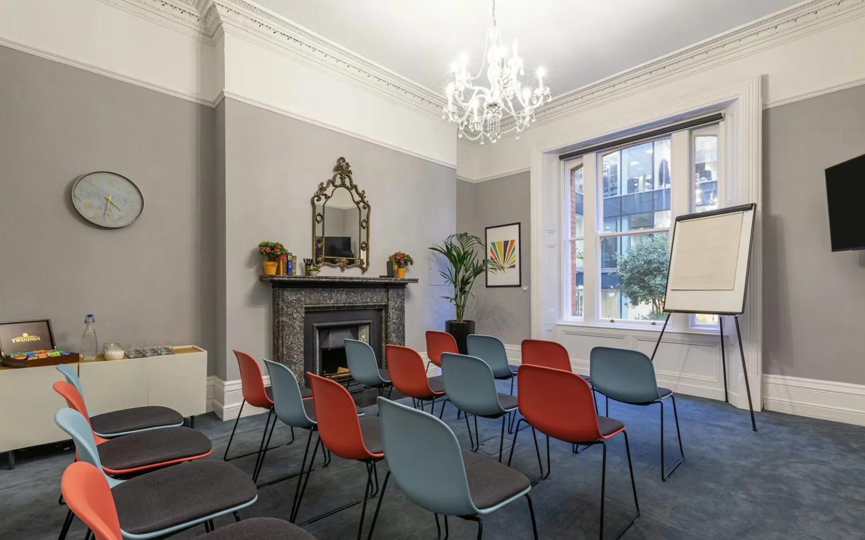 A conference room with chairs arranged in rows, a whiteboard on a stand, and a window with outdoor views. The room features a chandelier, fireplace, mirror, and artwork on the walls.