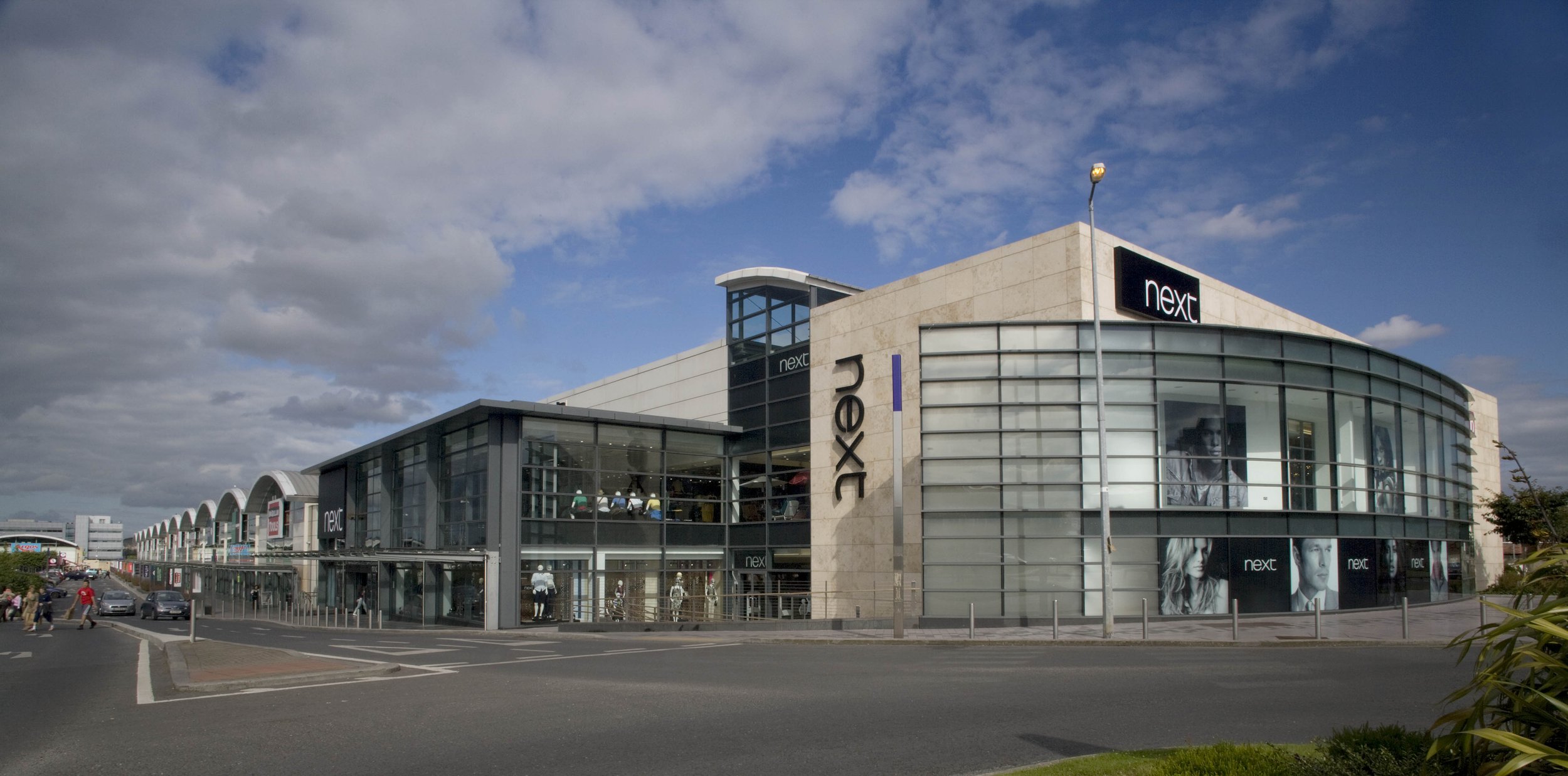 Large shopping mall building with signage for 'Next,' featuring glass exterior, modern architectural design, and a busy parking lot in front.