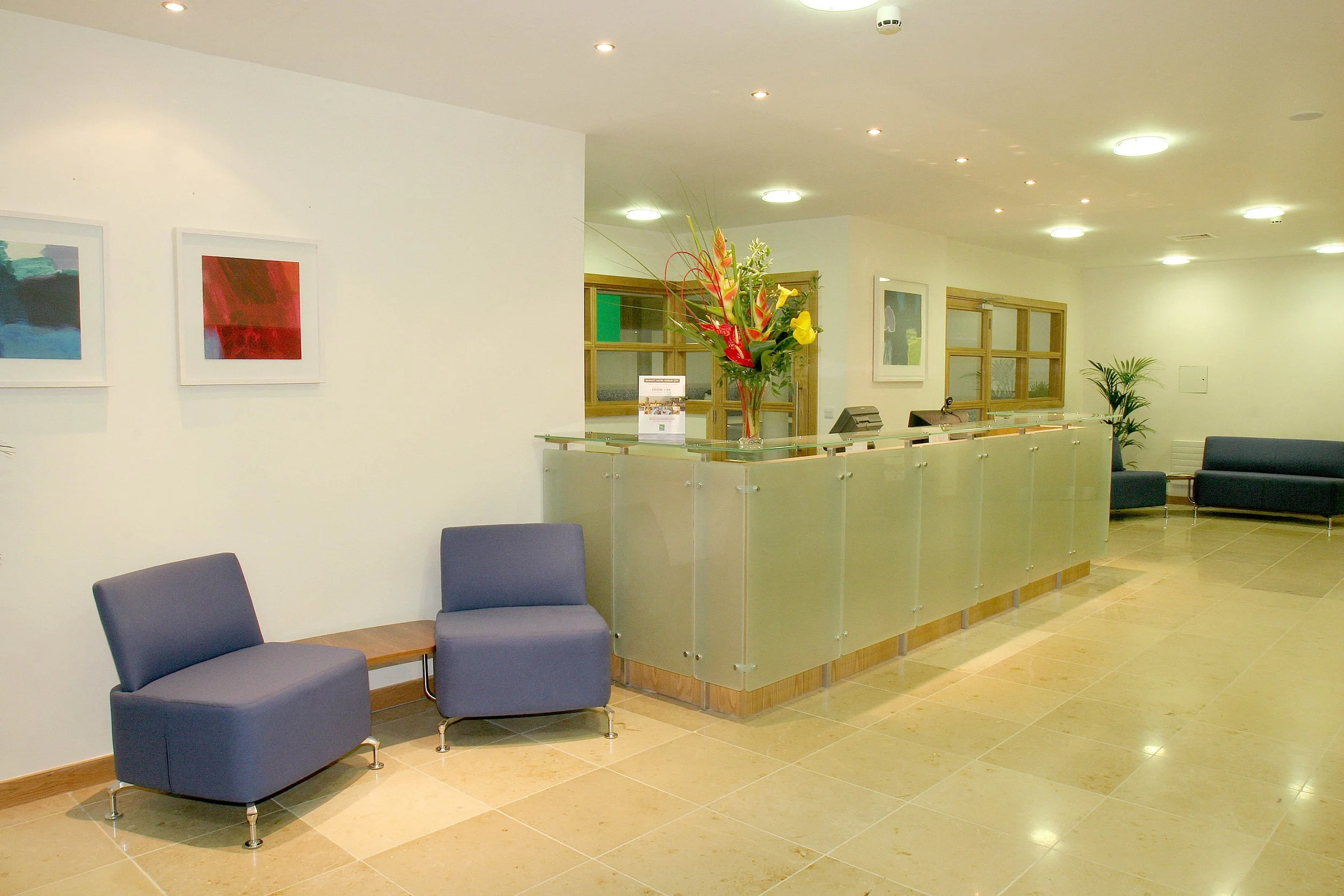 Hotel reception area with a glass counter, a large floral arrangement, seating area with blue chairs, framed artwork on the walls, and wooden accents.
