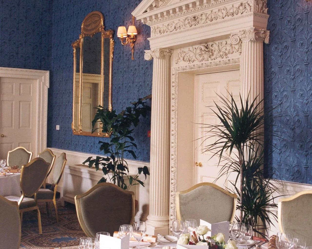 Elegant banquet room with blue patterned wallpaper, white ornate archway, and large mirror with gold frame. Several chairs around tables with place settings and floral centerpieces. Potted plants near walls and soft lighting.