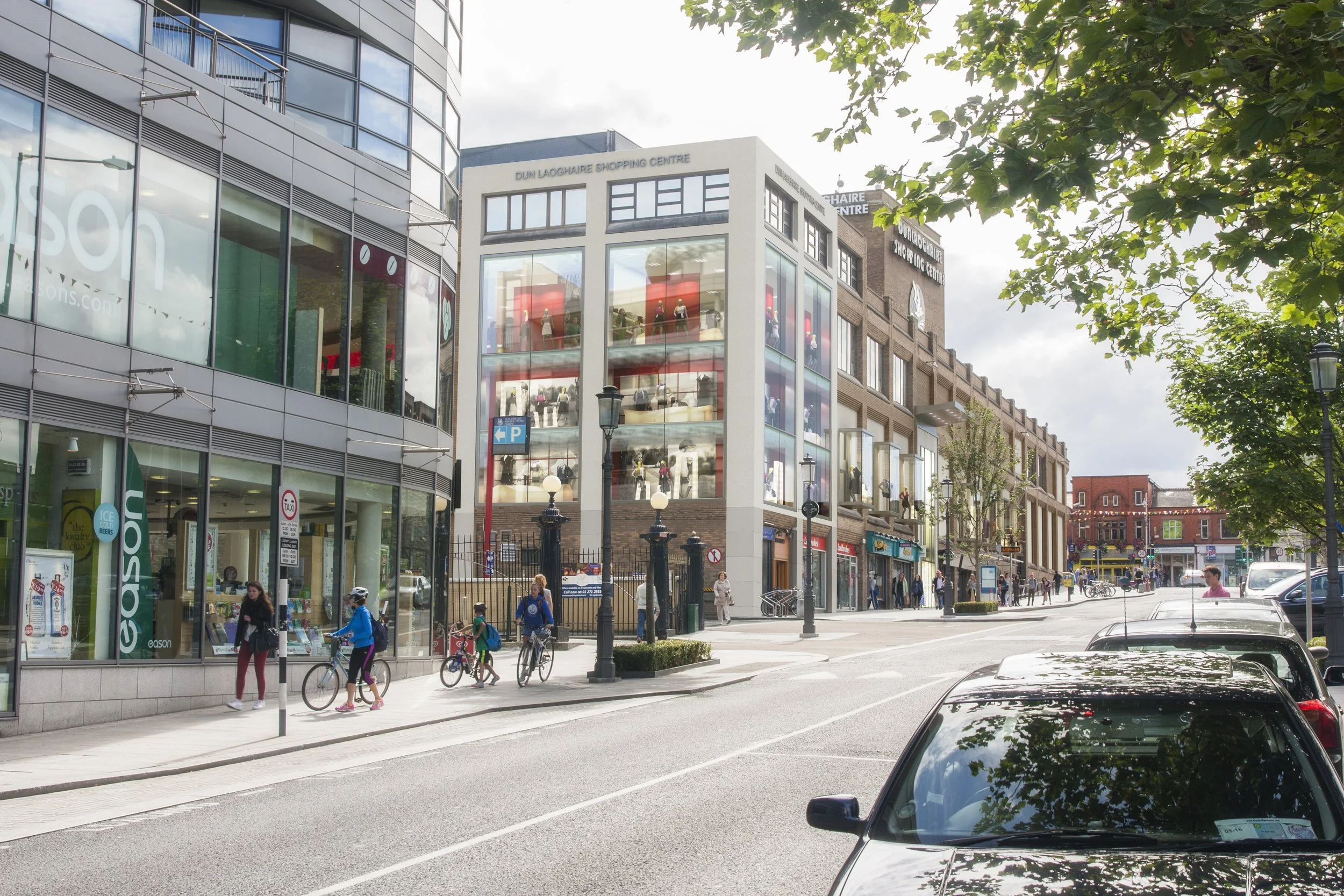 A busy city street with pedestrians, cyclists, parked cars, and modern commercial buildings, including a shopping center with large glass windows and store signs.