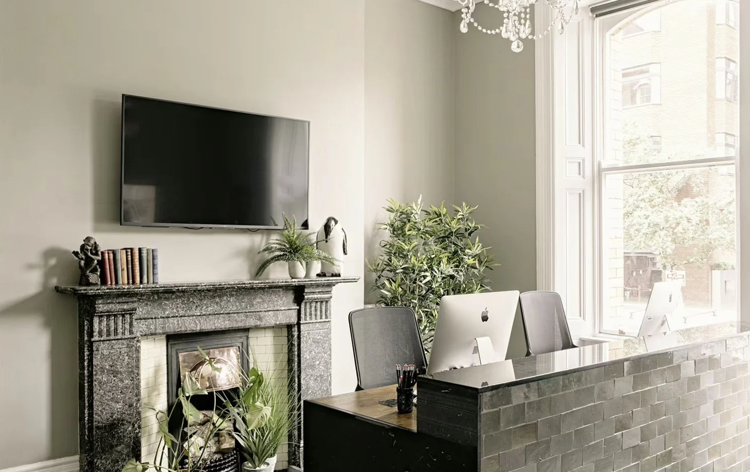 Modern office space with a black brick desk, computer monitors, a large window, potted plants, a framed picture, and a marble fireplace with decorative items above it.