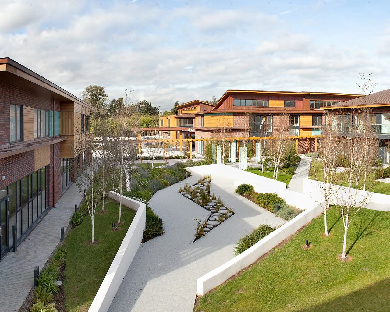 Modern residential buildings surrounding landscaped communal courtyard with pathways, small trees, and garden beds under a partly cloudy sky.