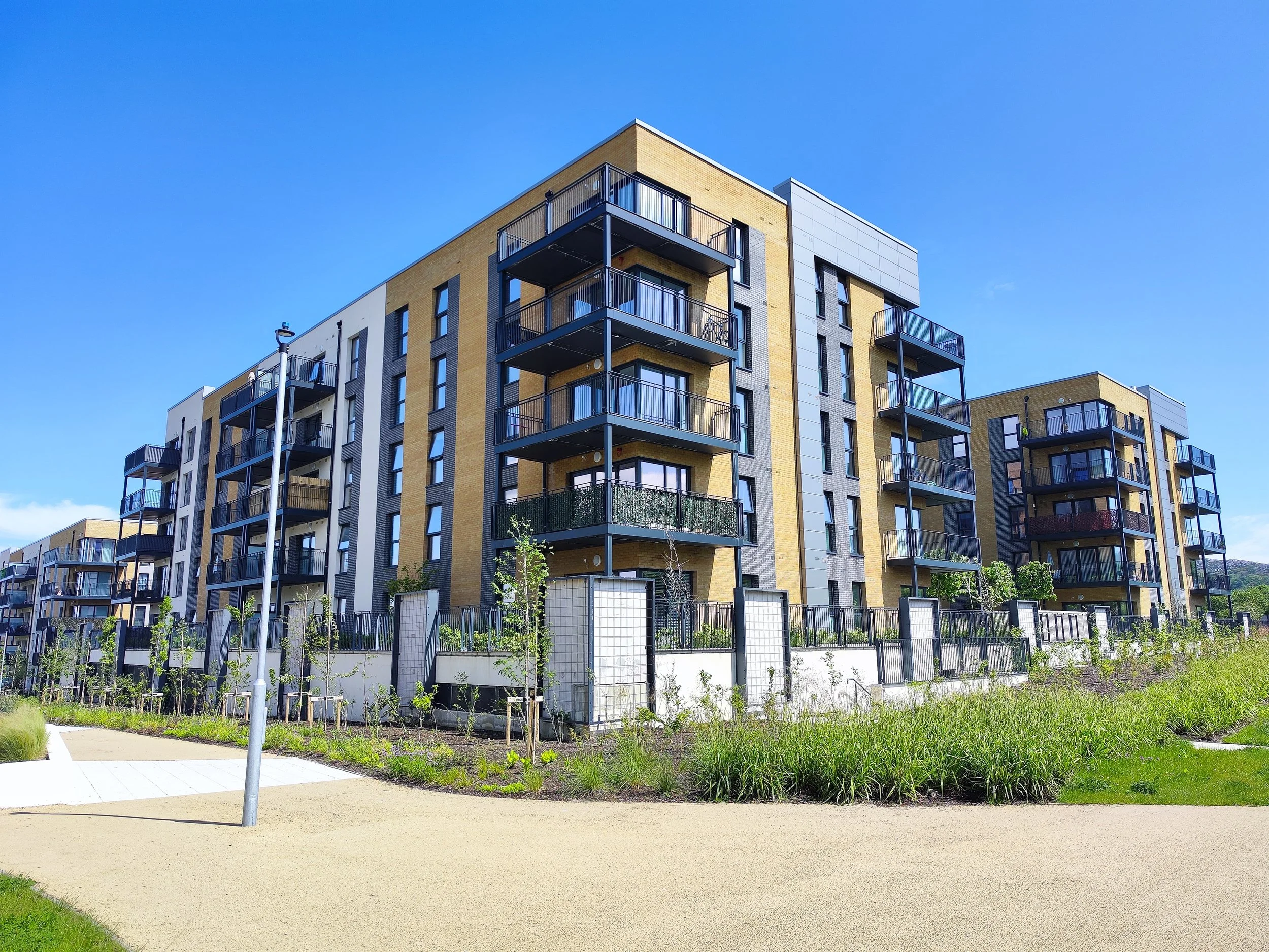 Modern multi-story apartment building with balconies, situated in a landscaped area with grass, small trees, and pathways under a clear blue sky.