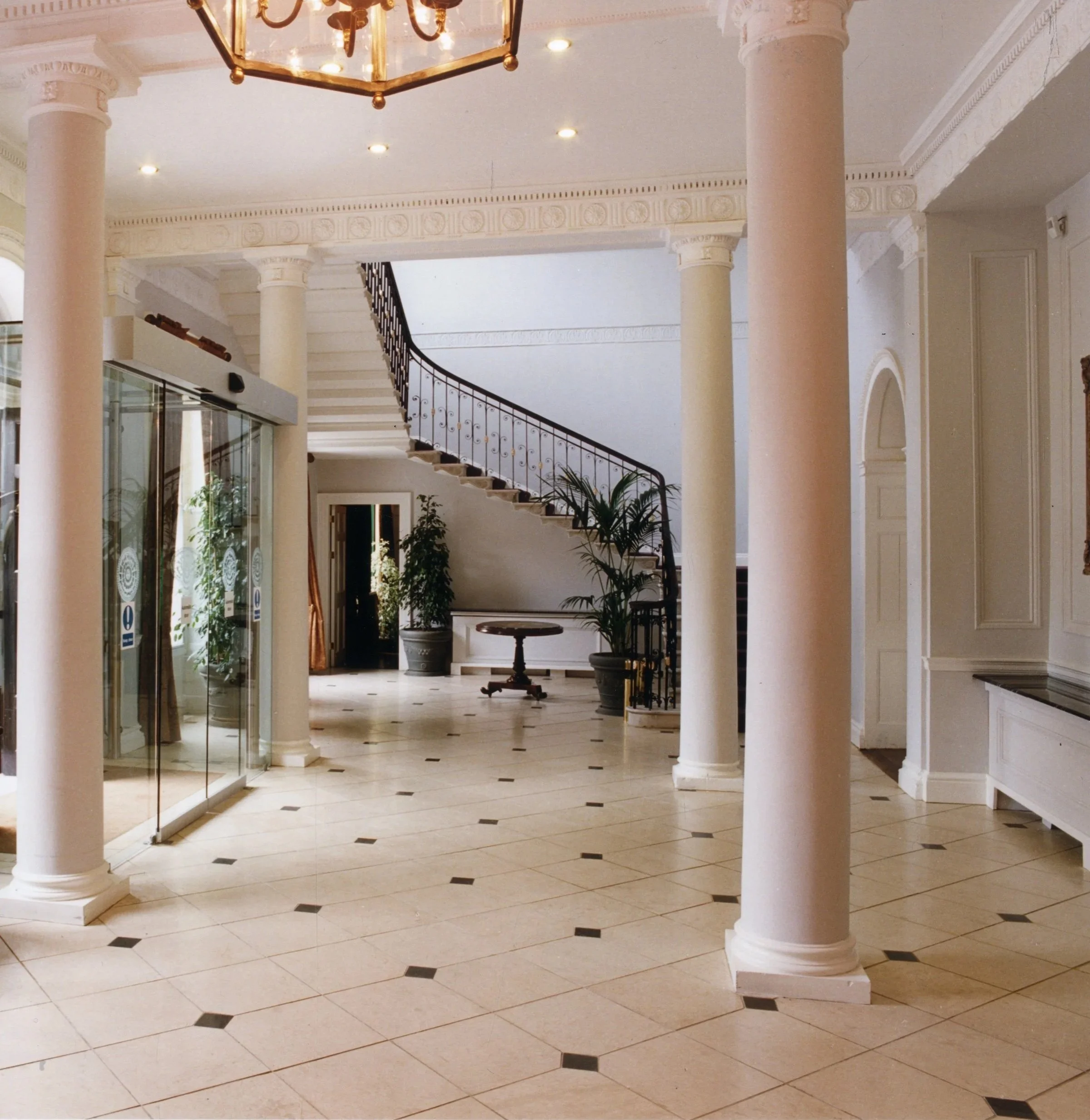 An elegant hotel lobby with cream-colored tiled floor, white pillars, and a staircase with black railing leading upstairs. There are potted plants near the entrance and a round wooden table in the background.