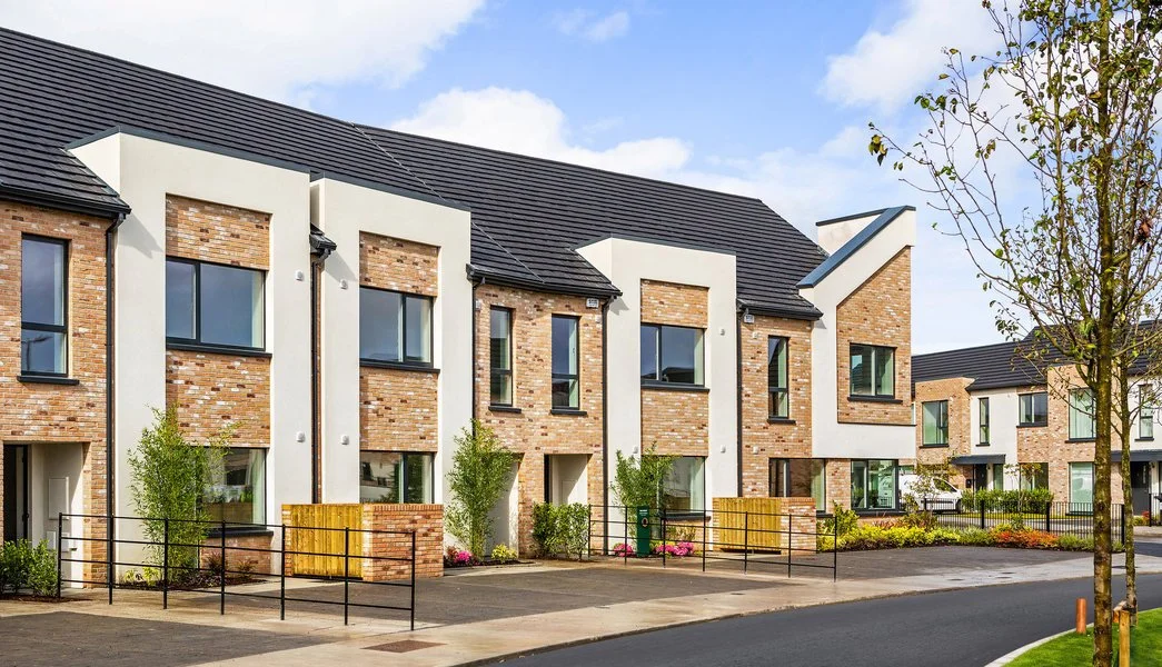 Modern residential apartment complex with brick and white exterior, multiple windows, small front yards with trees, and a paved walkway