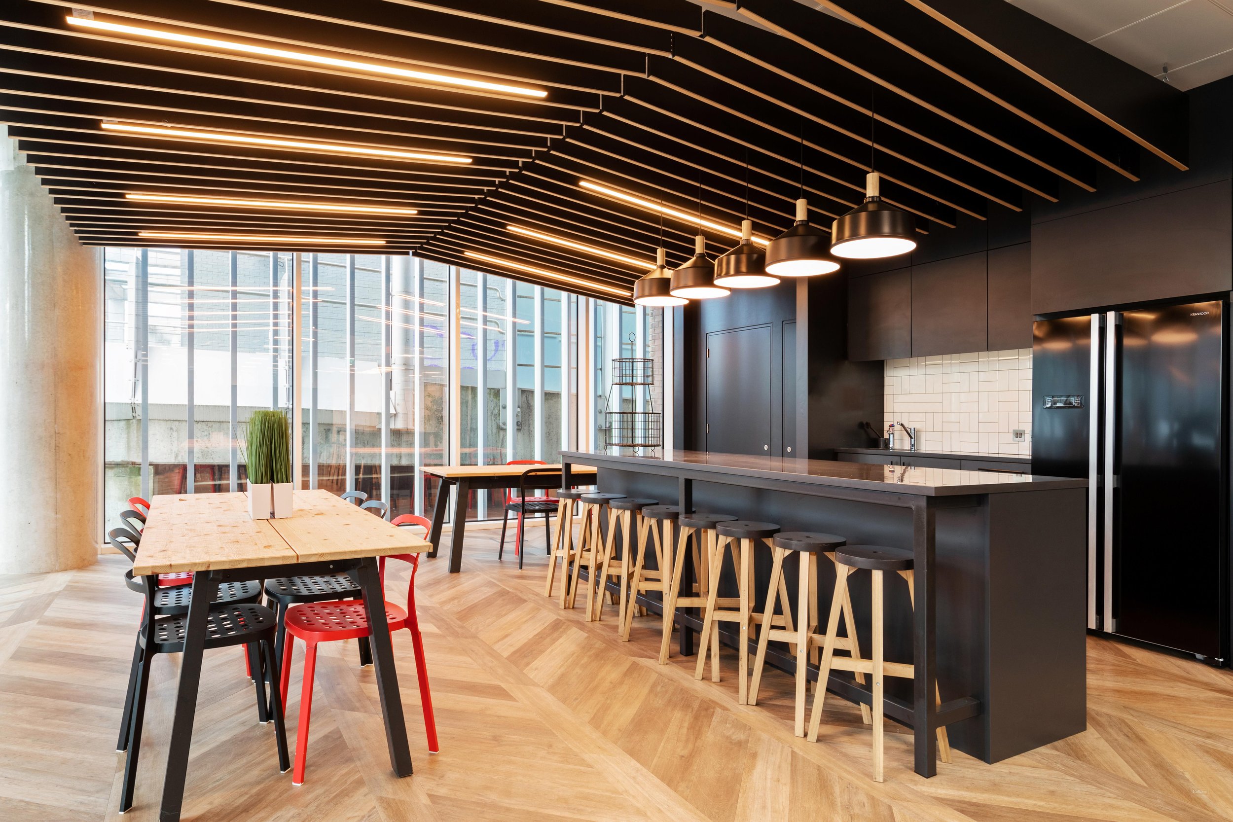 Modern kitchen and dining area with wooden floors, black and red chairs, a large wooden table with a flower vase, a black bar counter with black stools, pendant lights, and large glass windows.
