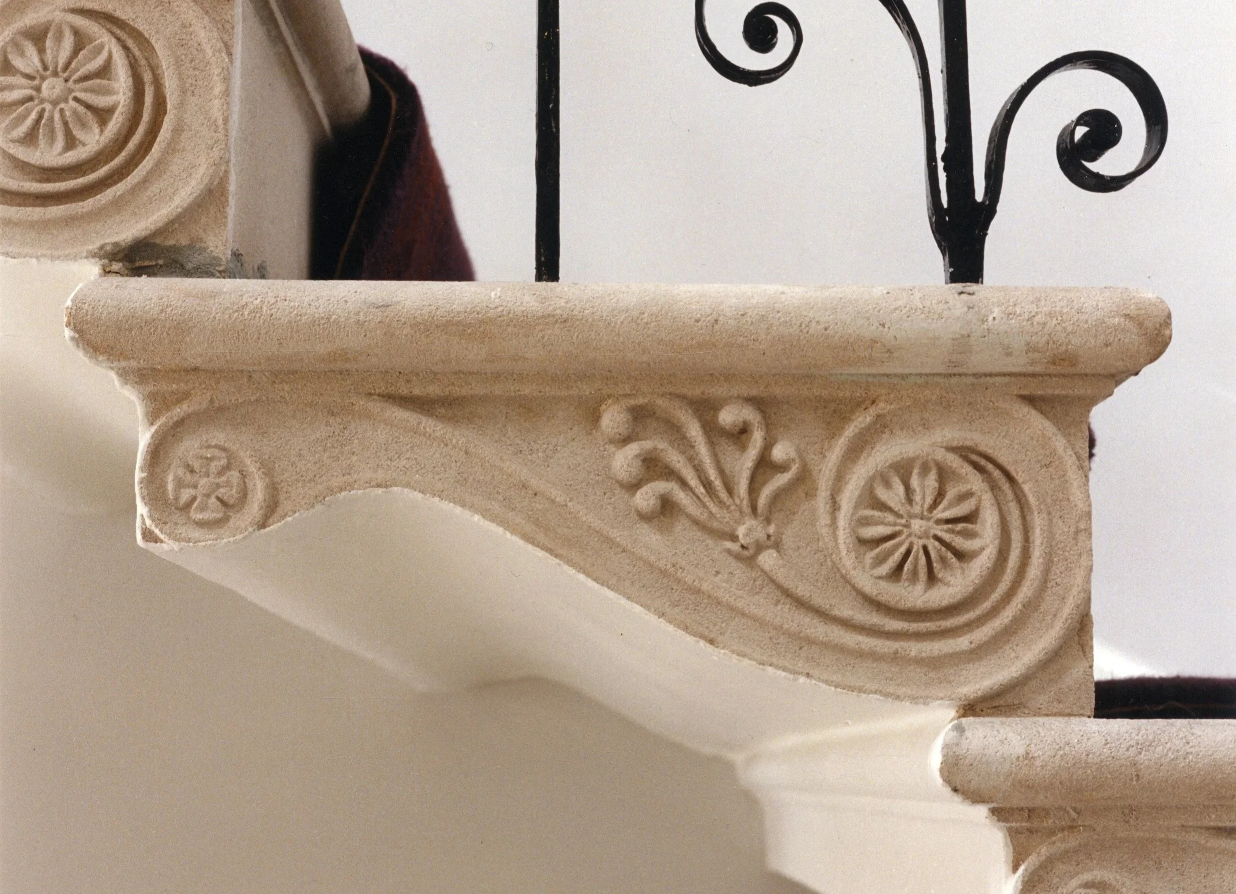 Close-up of decorative stone corbel with floral and scroll carvings supporting a shelf, with part of a wrought iron rail and wall visible in background.