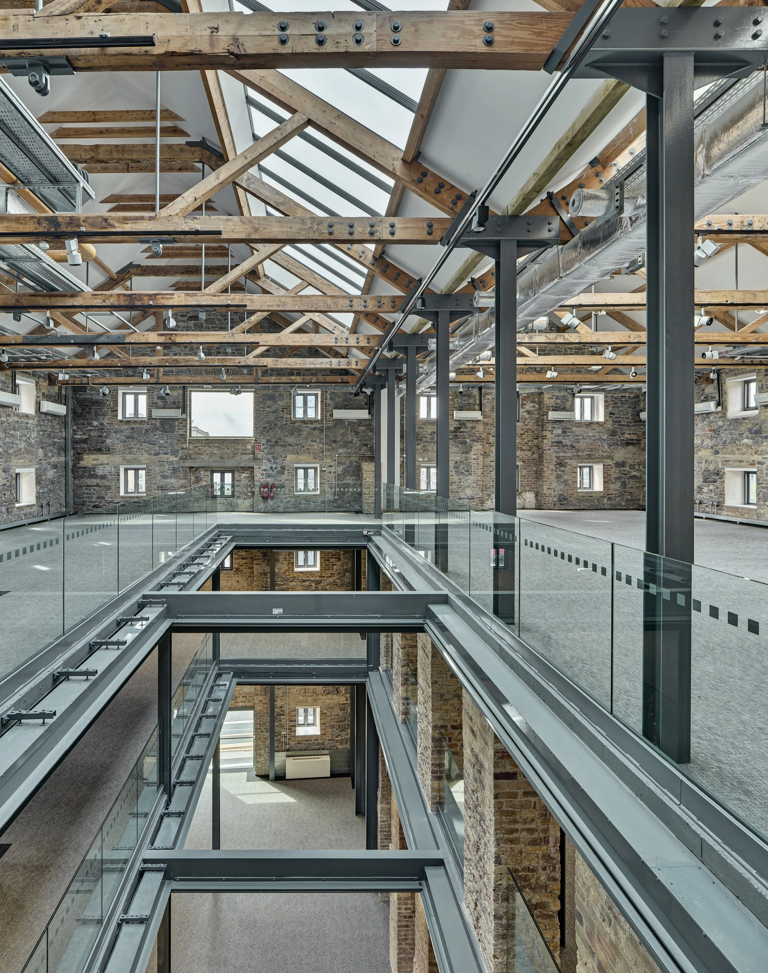 Interior view of a modern building with an open atrium, exposed wooden beams on the ceiling, brick walls with small windows, and glass railing walkways.
