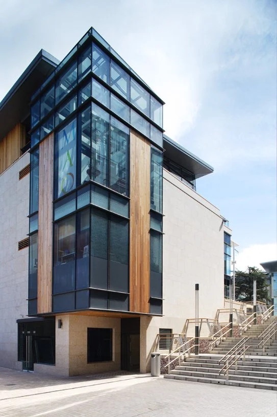 Modern multi-story building with large glass windows and wooden paneling, with stairs leading up to the entrance.