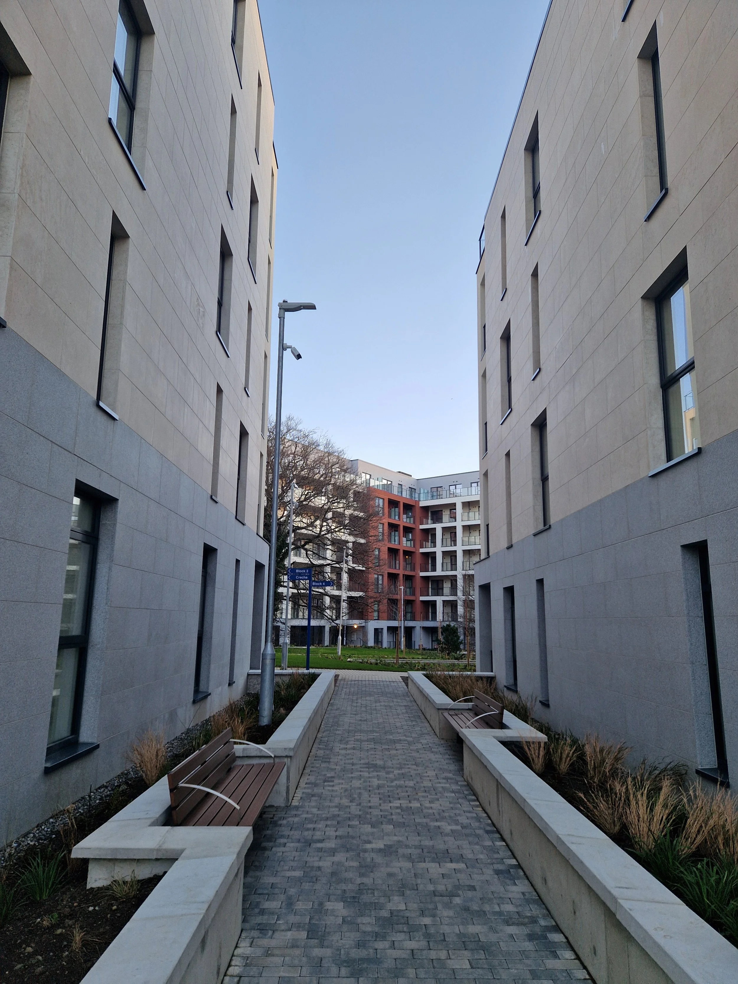 A paved walkway flanked by two modern apartment buildings with beige and gray facades, outdoor benches, and landscaped areas, leading to additional apartment buildings in the background.