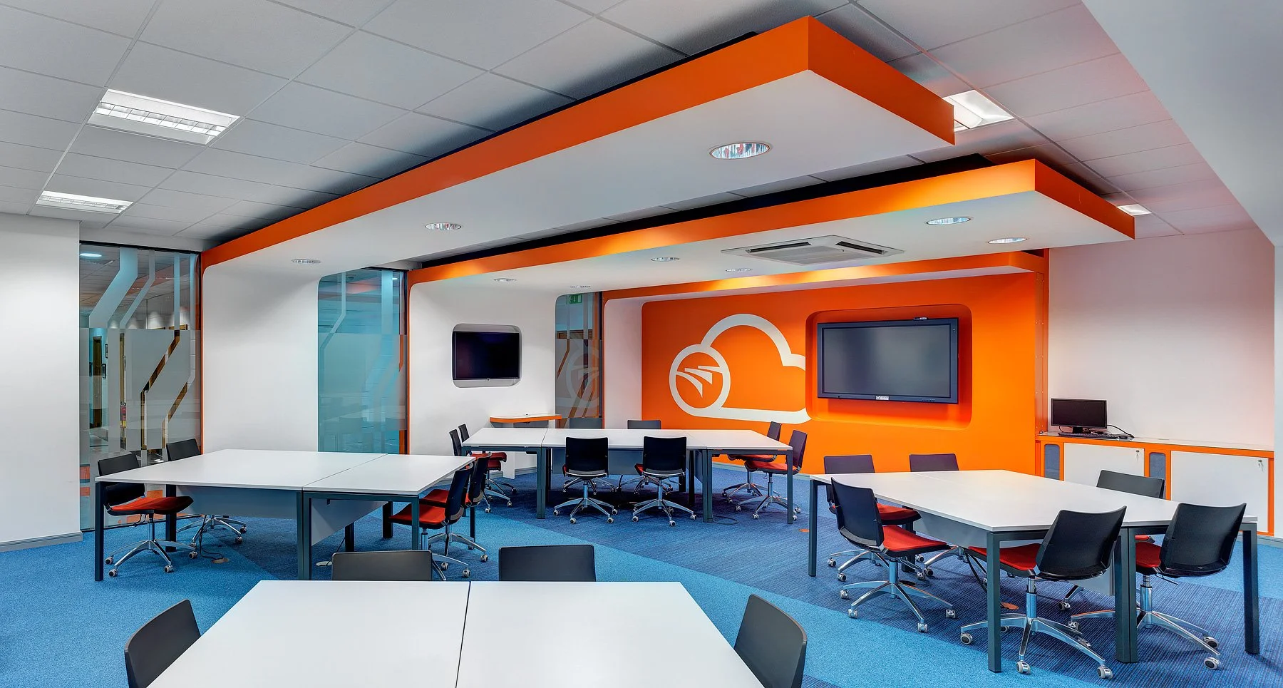 Modern conference room with white tables, black and orange chairs, large TV screens, and a prominent orange and white logo on the back wall.