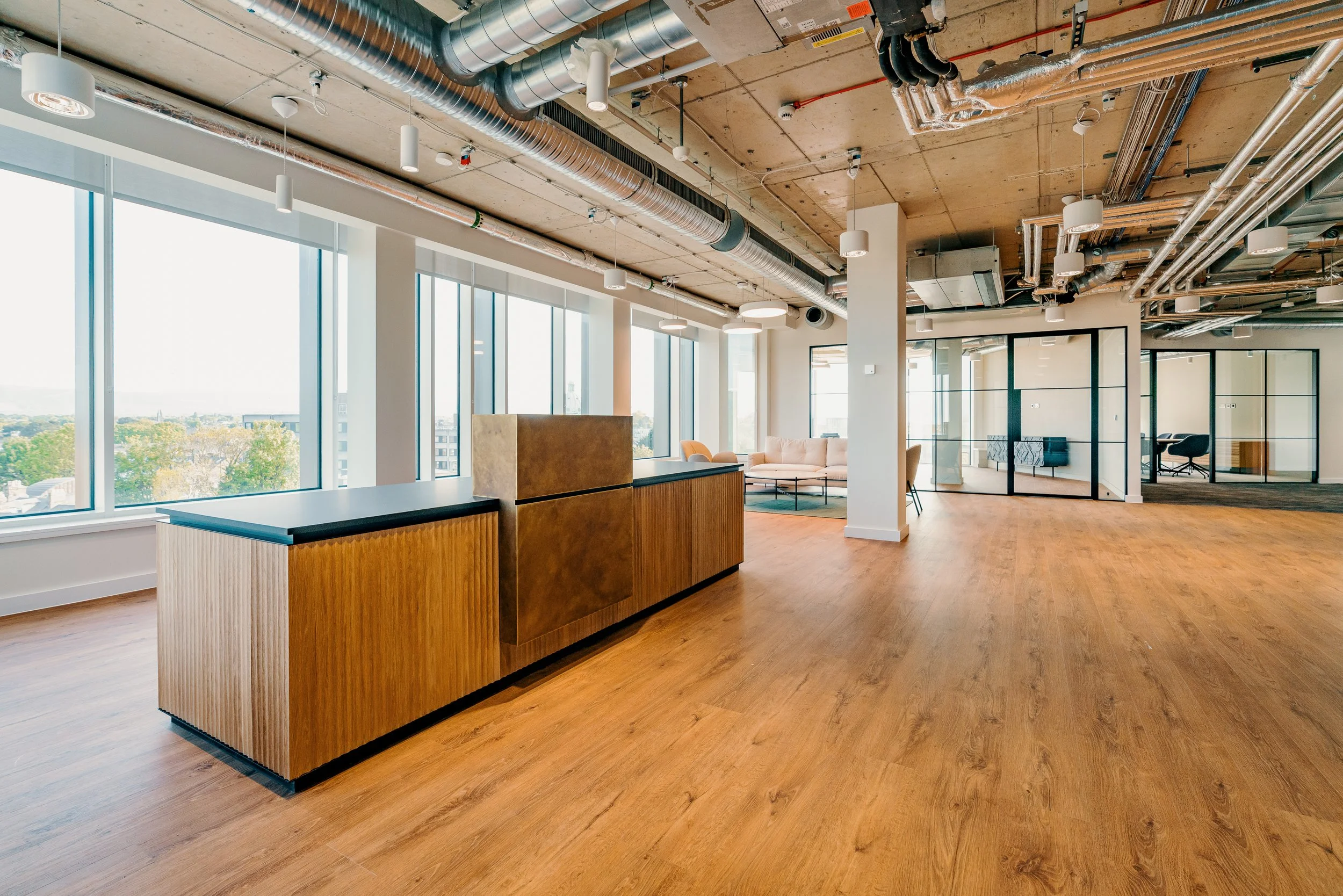 Modern office space with large windows, wooden flooring, a reception desk, and a sitting area with sofas and chairs, exposed ceiling pipes, and glass-walled conference rooms.