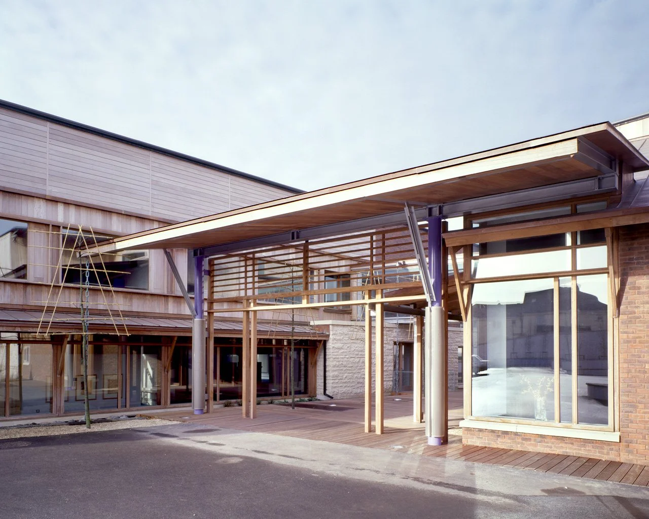 Modern building with wooden and glass exterior, featuring a large glass window and a covered entrance supported by metal beams.
