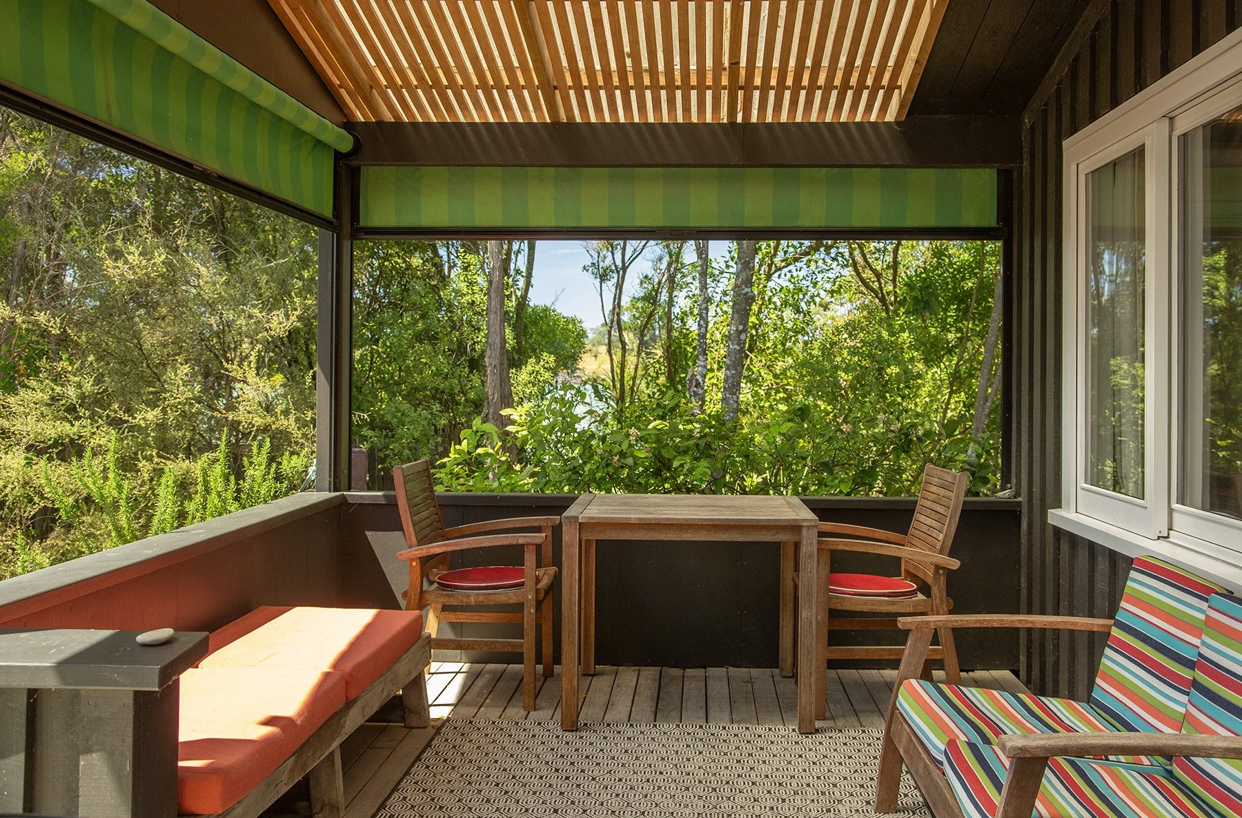 A screened porch with wooden furniture, including a table, chairs with red cushions, a striped chair, and an orange cushioned bench, overlooking lush greenery and trees.