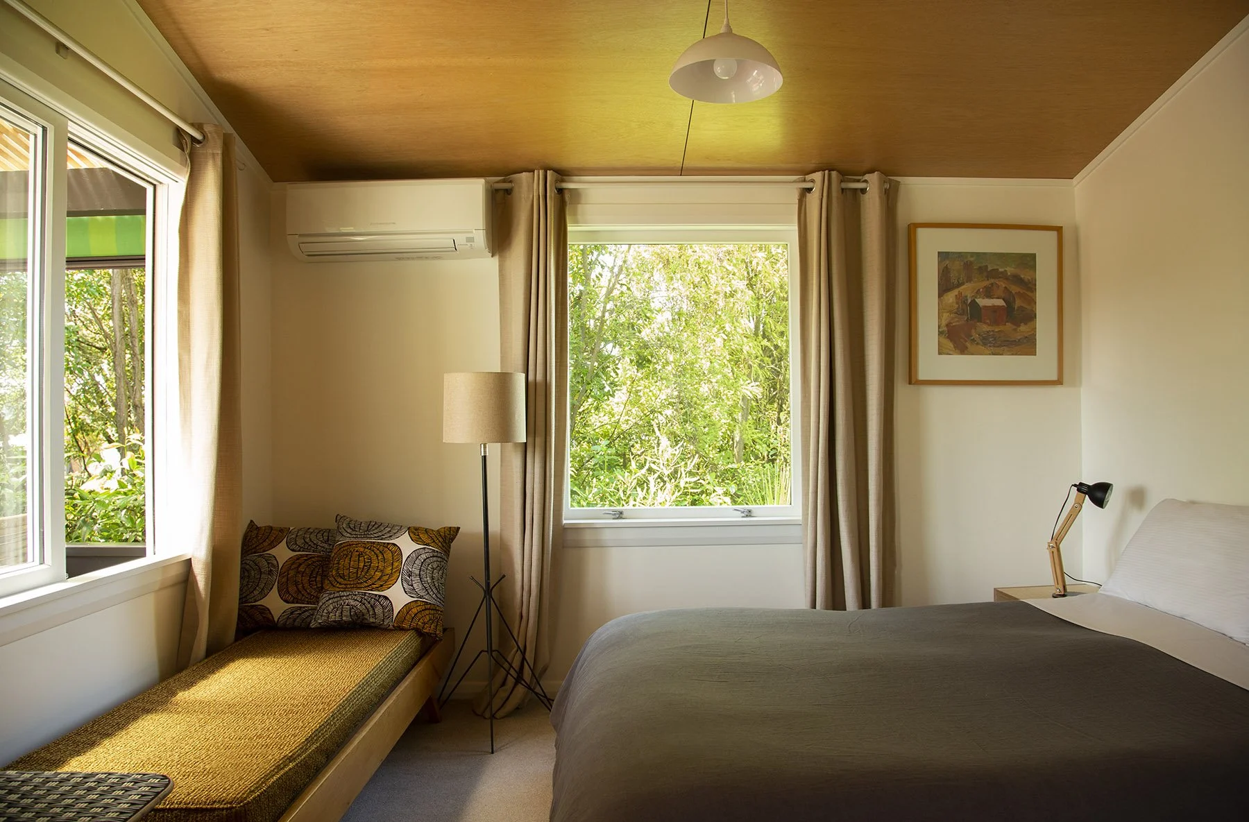 A cozy bedroom with two windows, beige curtains, a bed with a dark comforter, a floor lamp, a painting on the wall, and greenery outside the windows.