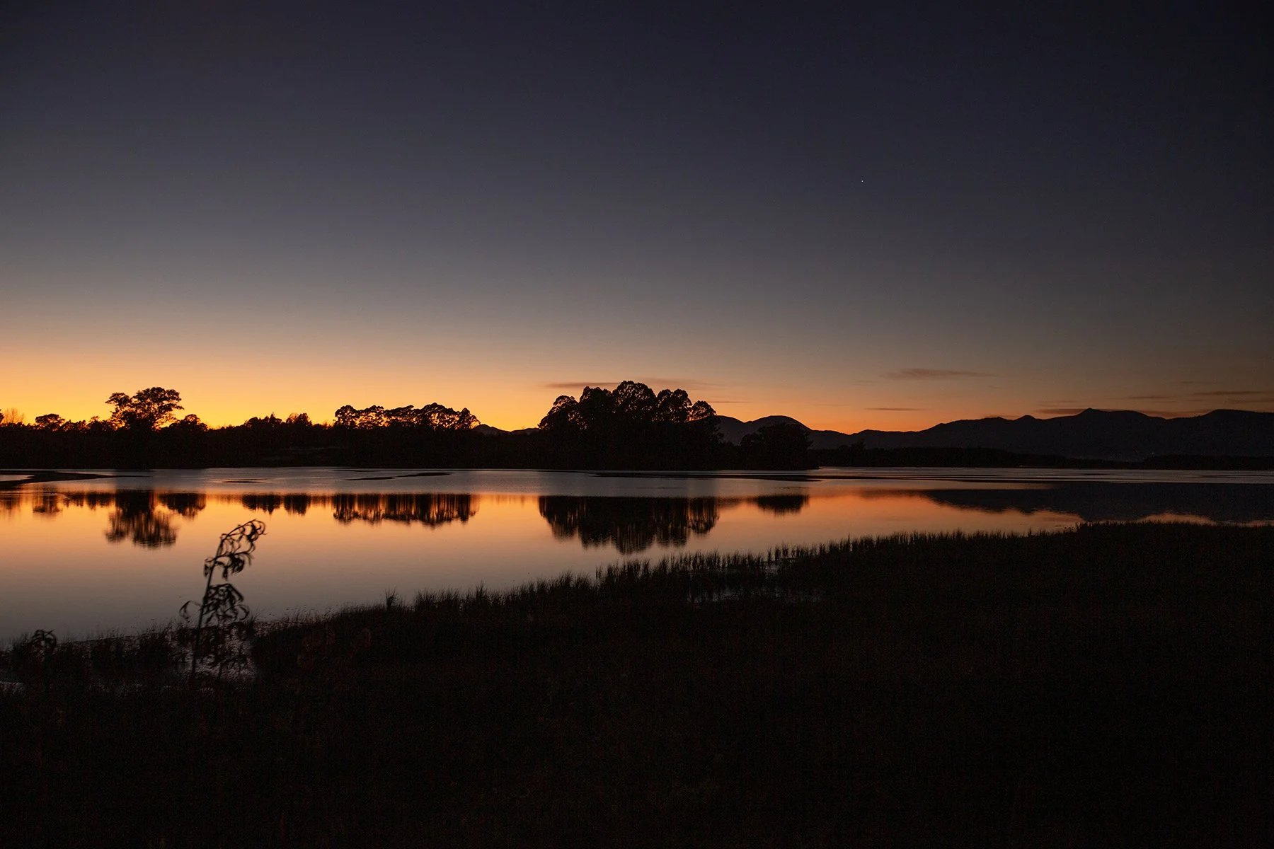 Serene sunset over a calm inlet with silhouettes of trees and distant mountains reflected in the water.