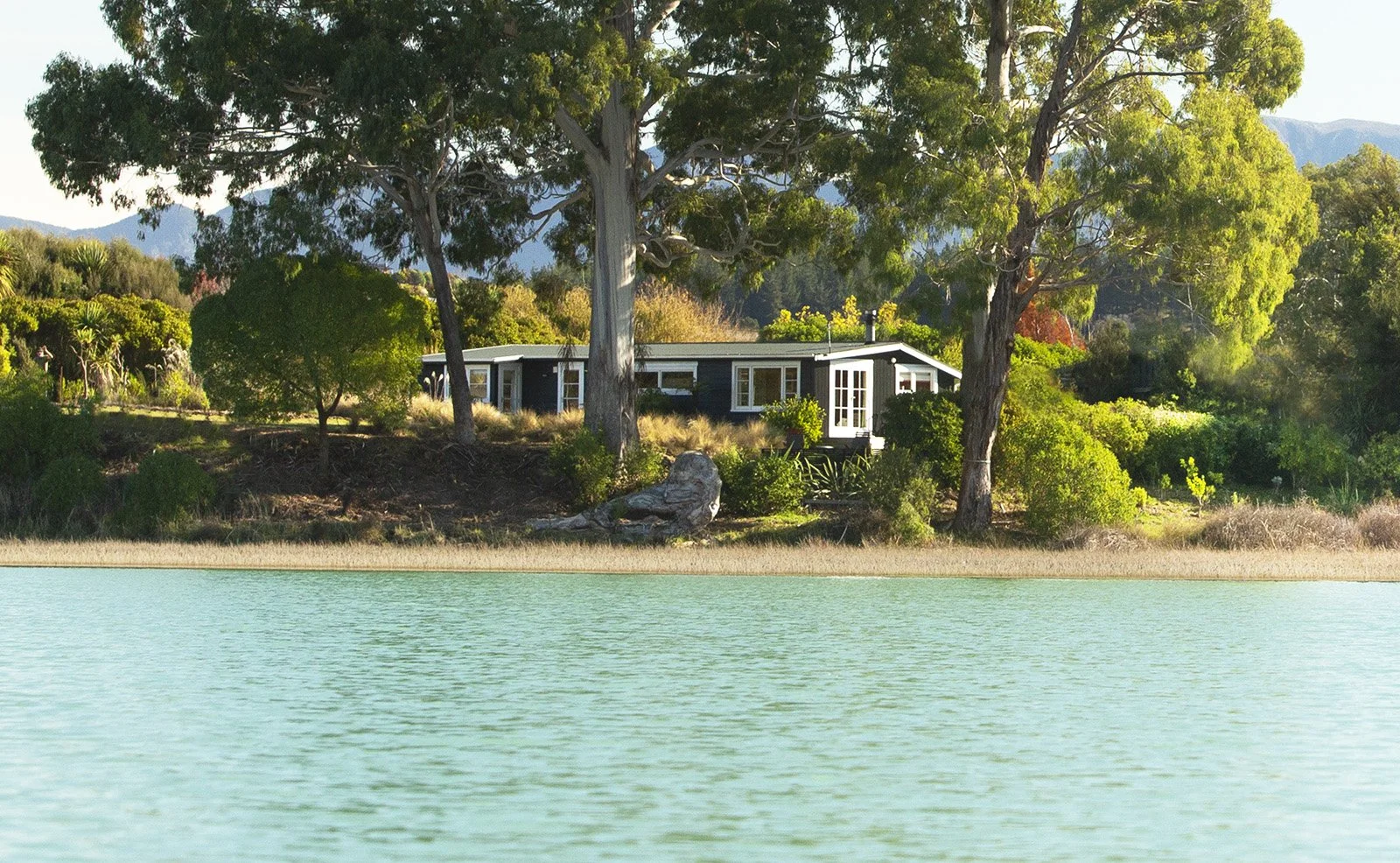 Warm-lit black cabin by water with mountain backdrop — Mapua accommodation