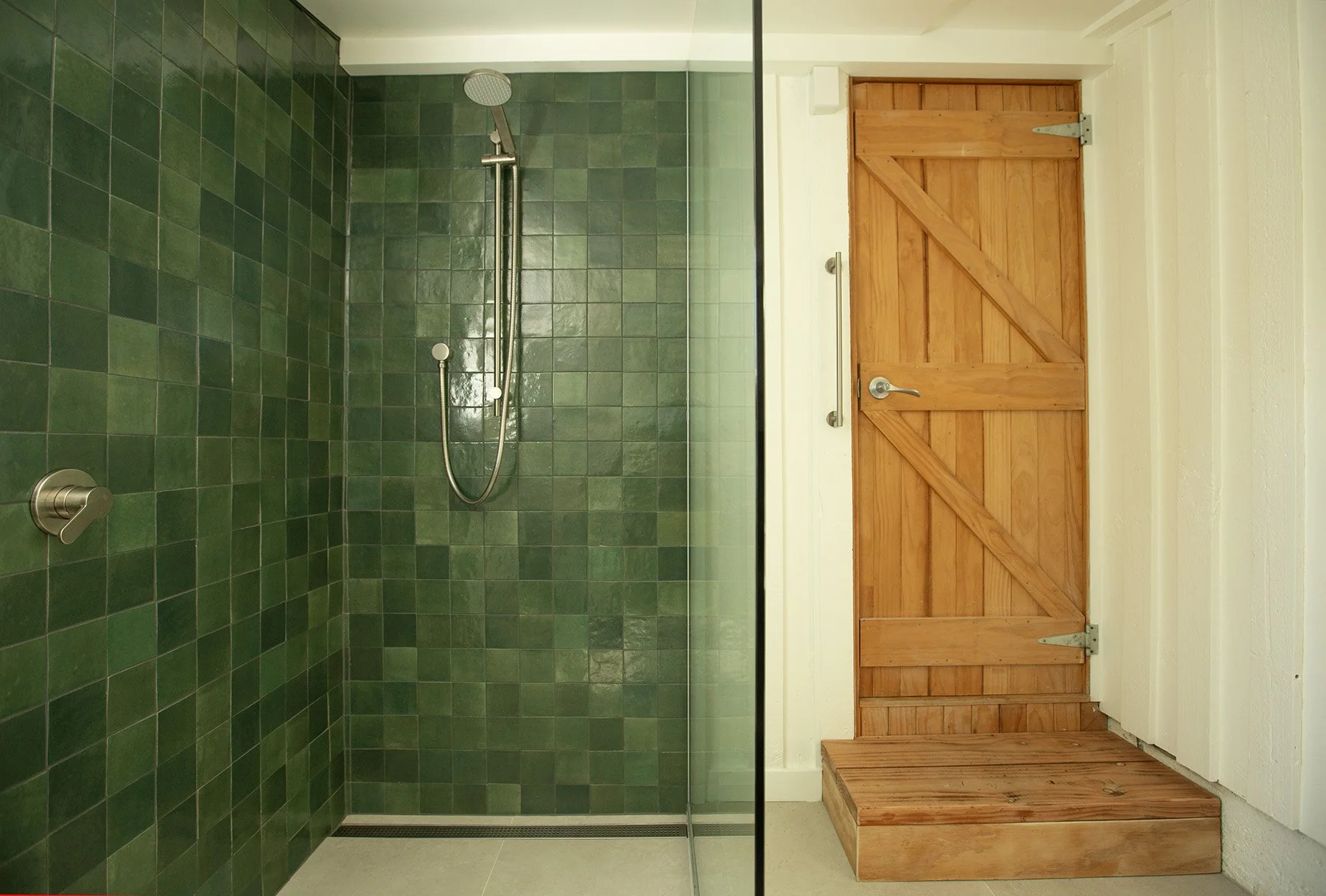 A modern shower area with green tiled walls, a chrome shower head, and a glass partition, next to a wooden door with a latch and a small platform at the base.