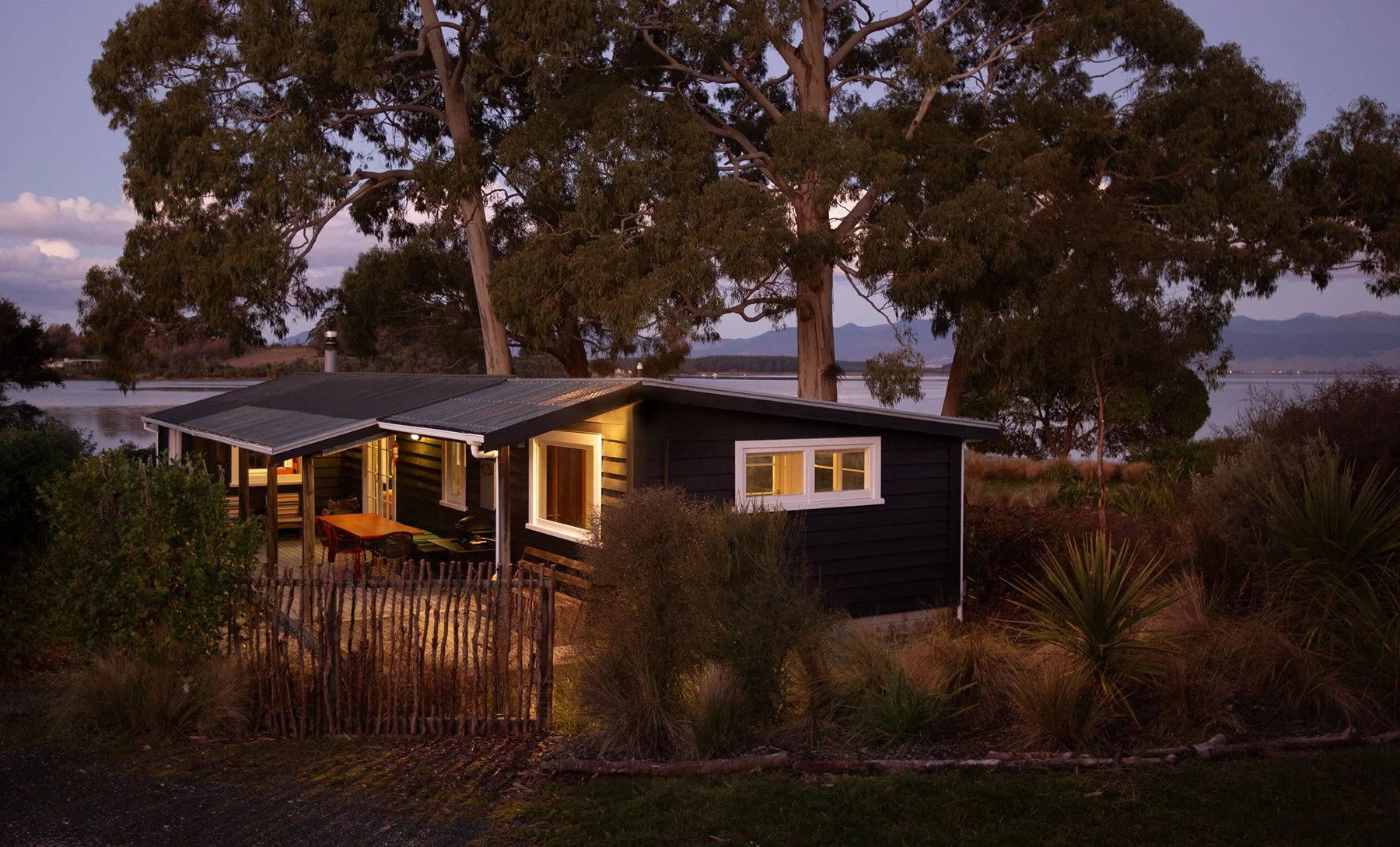 A small black house with a metal roof seen at dusk, surrounded by trees and bushes, with a tidal inlet and mountains in the background.