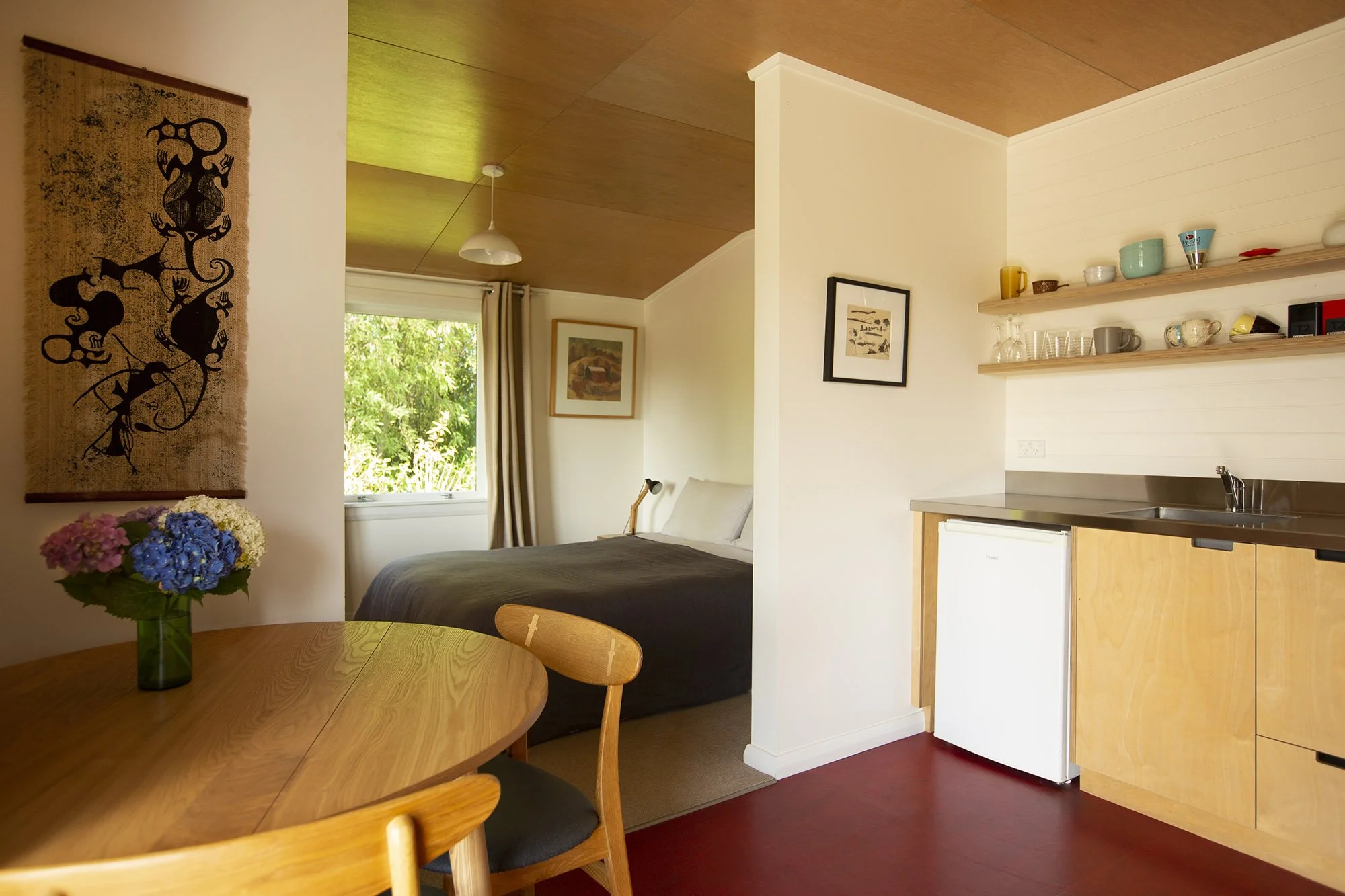 Interior view of a cozy studio with a dining table, a small kitchenette with open shelves, a bed in the background, and a window showing greenery outside.