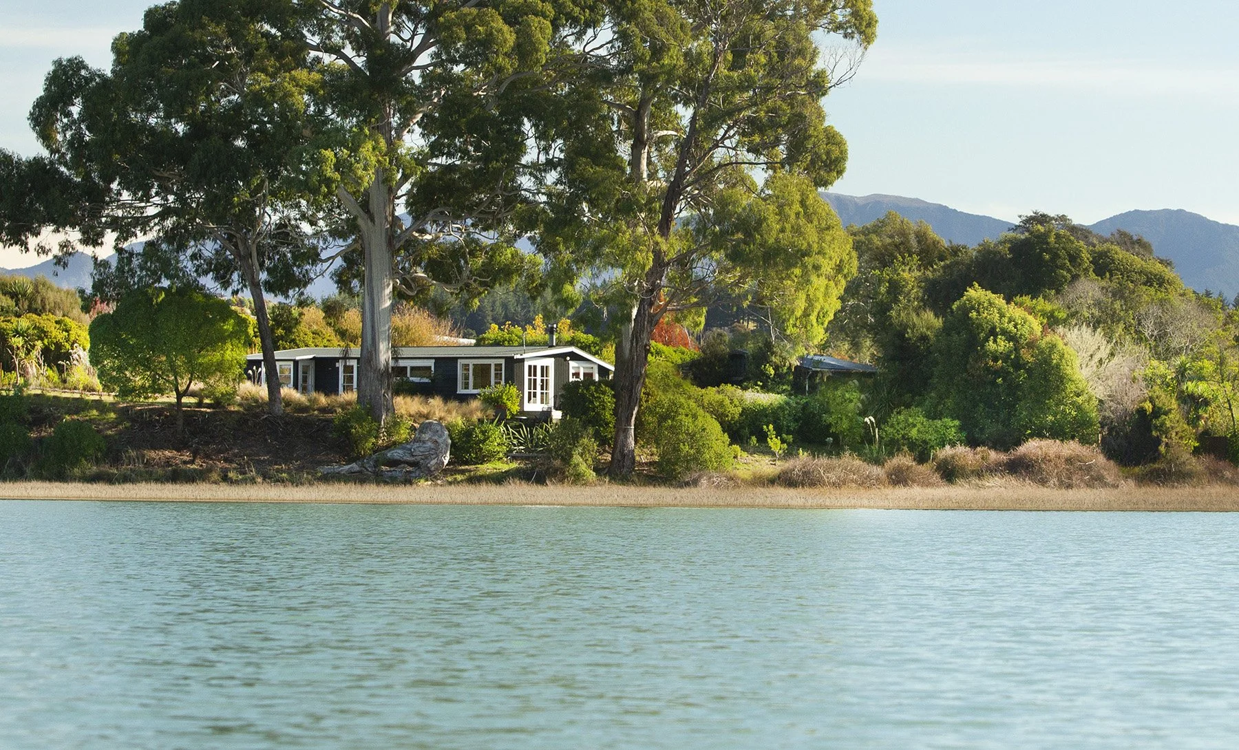 Crystal blue water of the Waimea inlet estuary surrounds the exterior of Karaka Cottage, which is a black and white original 50's Apple Pickers' Cottage. The cottage is surrounded by large gum trees with bright green foliage, and a range of other shr