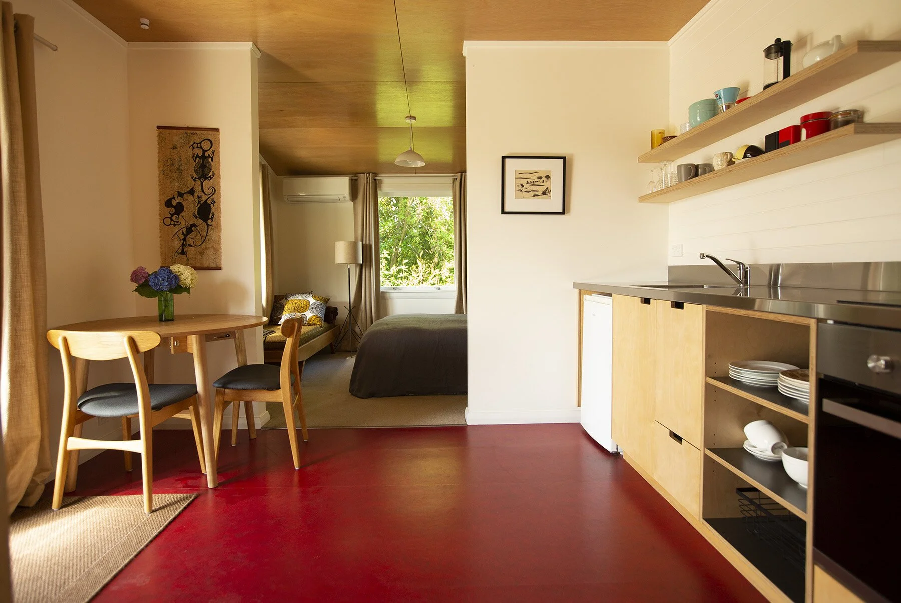 Open-concept living space with a dining area, kitchenette, and bedroom. Wooden dining table with four chairs, a small kitchen with open shelves holding cups and plates, a sink, and a small refrigerator. The bedroom has a bed, a window with curtains,