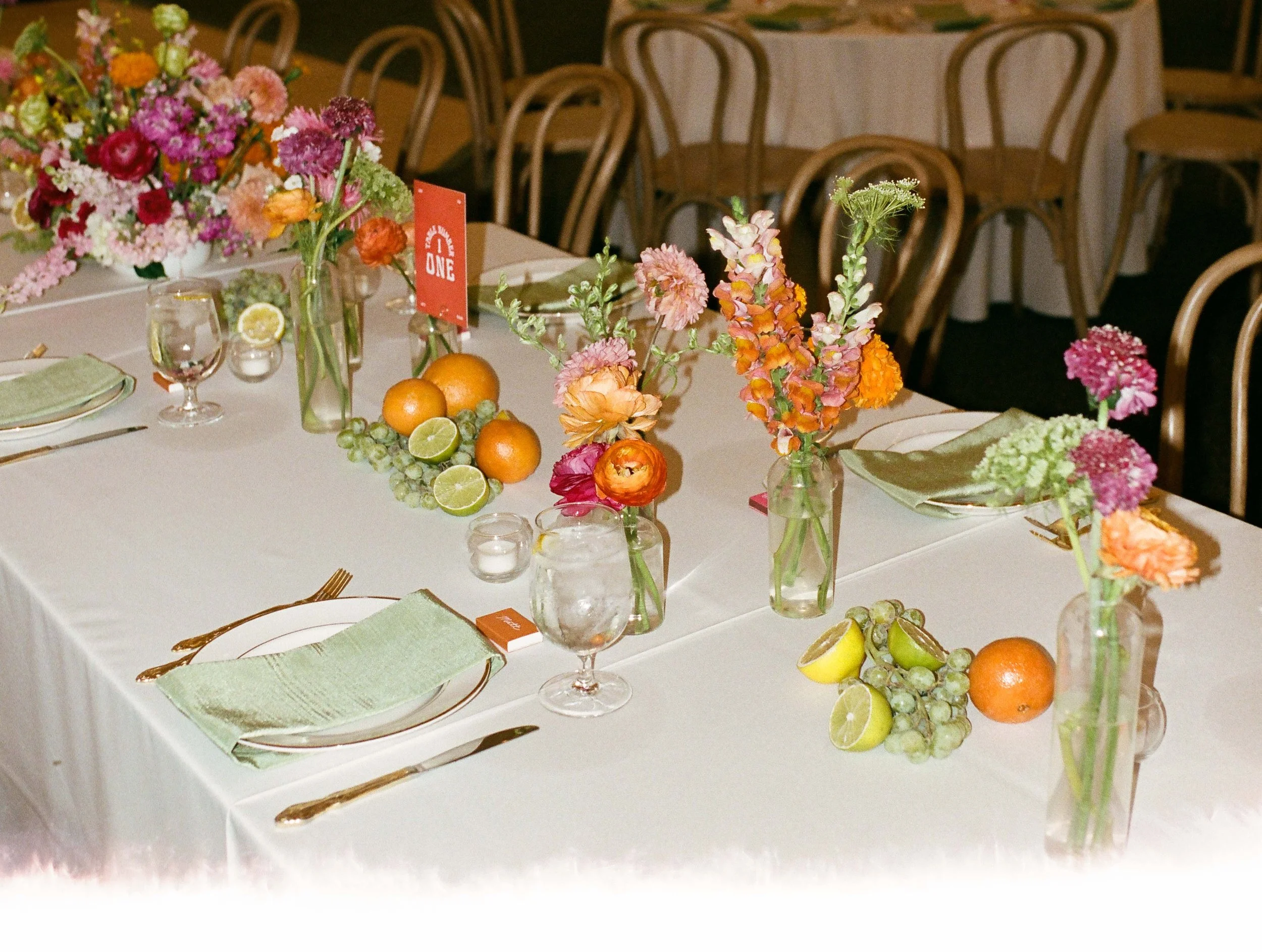 Wedding reception table with citrus, grapes, and bud vases filled with pink and orange flowers
