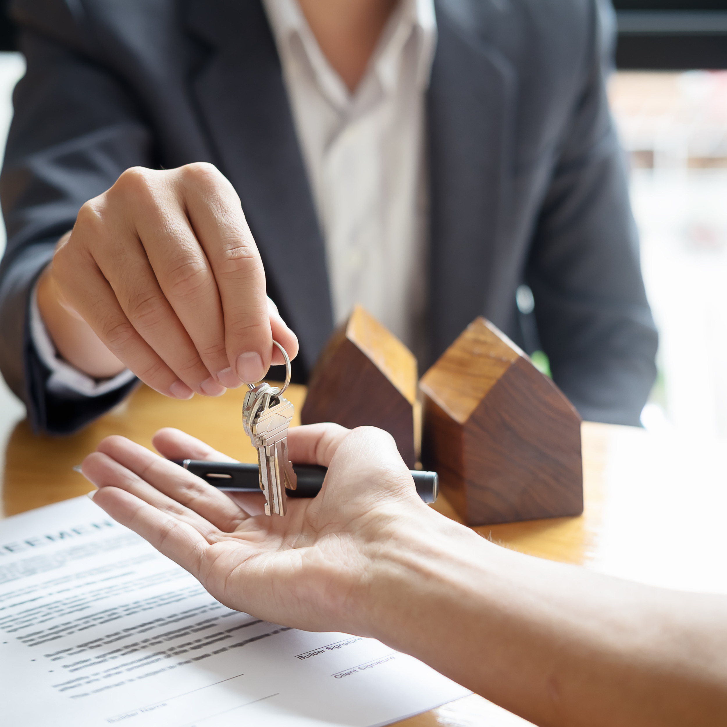 Person handing over house keys to another person across a table with contract and wooden house models.