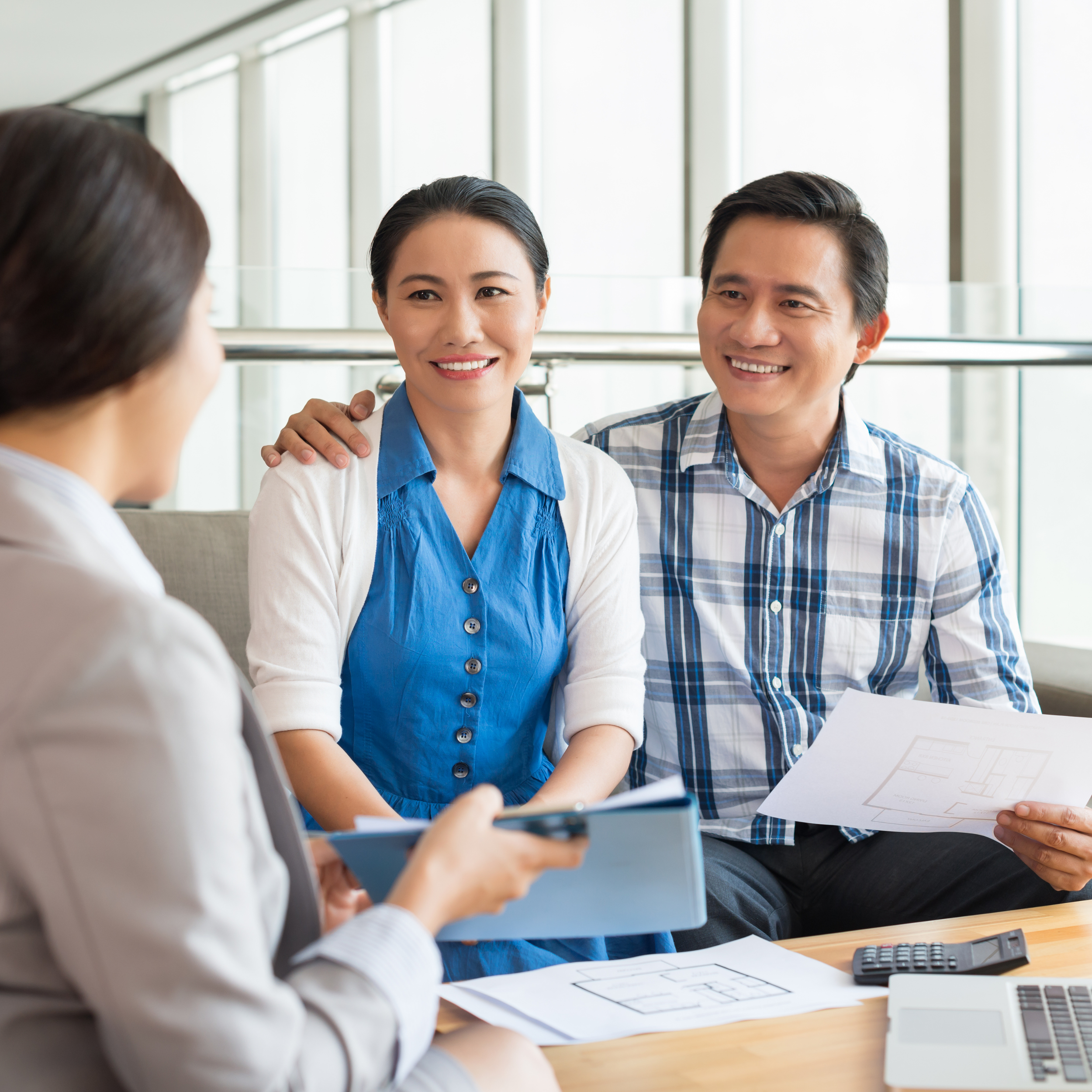 Couple discussing paperwork with a businesswoman at a table, holding documents and a blueprint, with a calculator and laptop nearby.