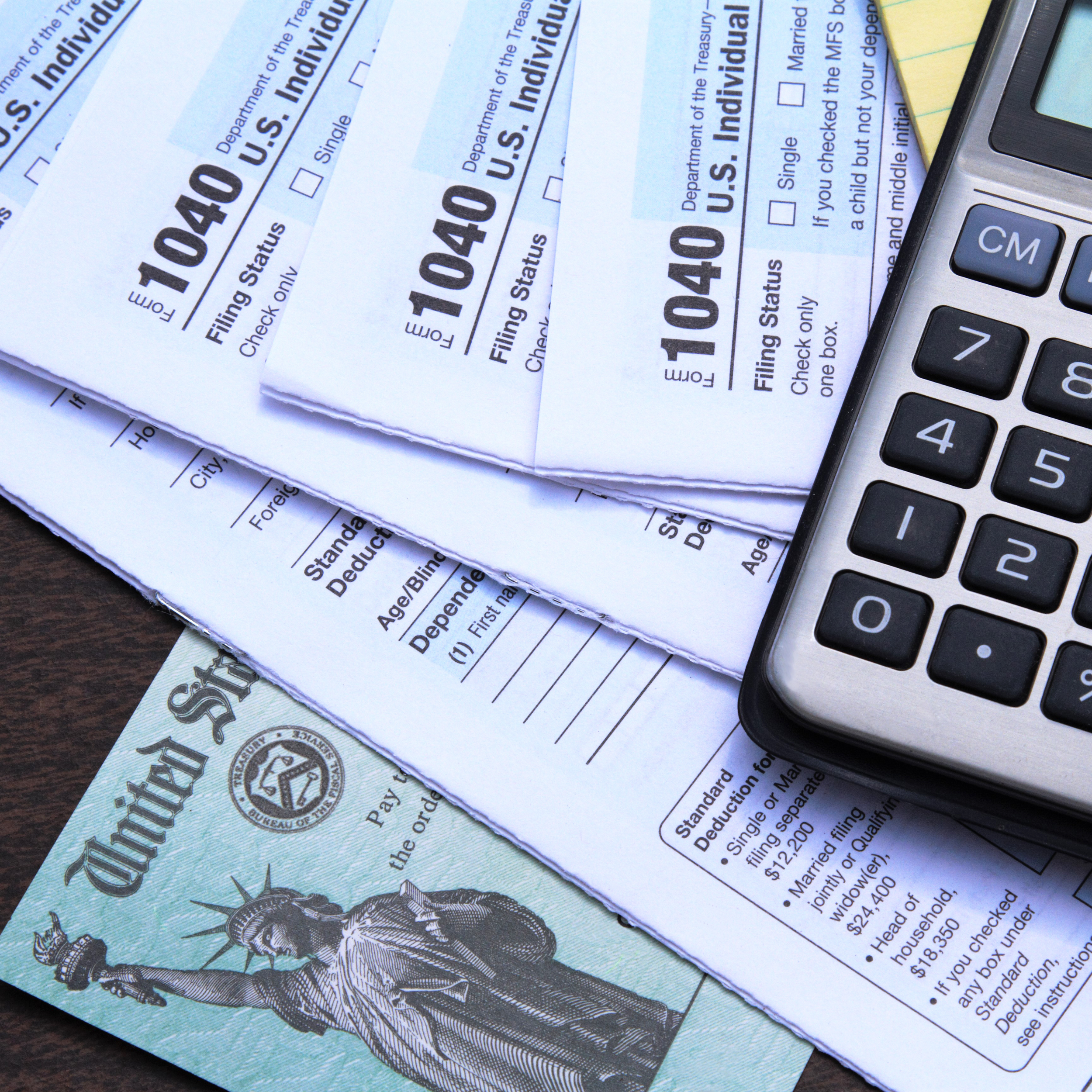 Close-up of U.S. tax forms 1040 atop a wooden desk with a calculator and a U.S. Treasury check, indicating tax preparation and financial planning.