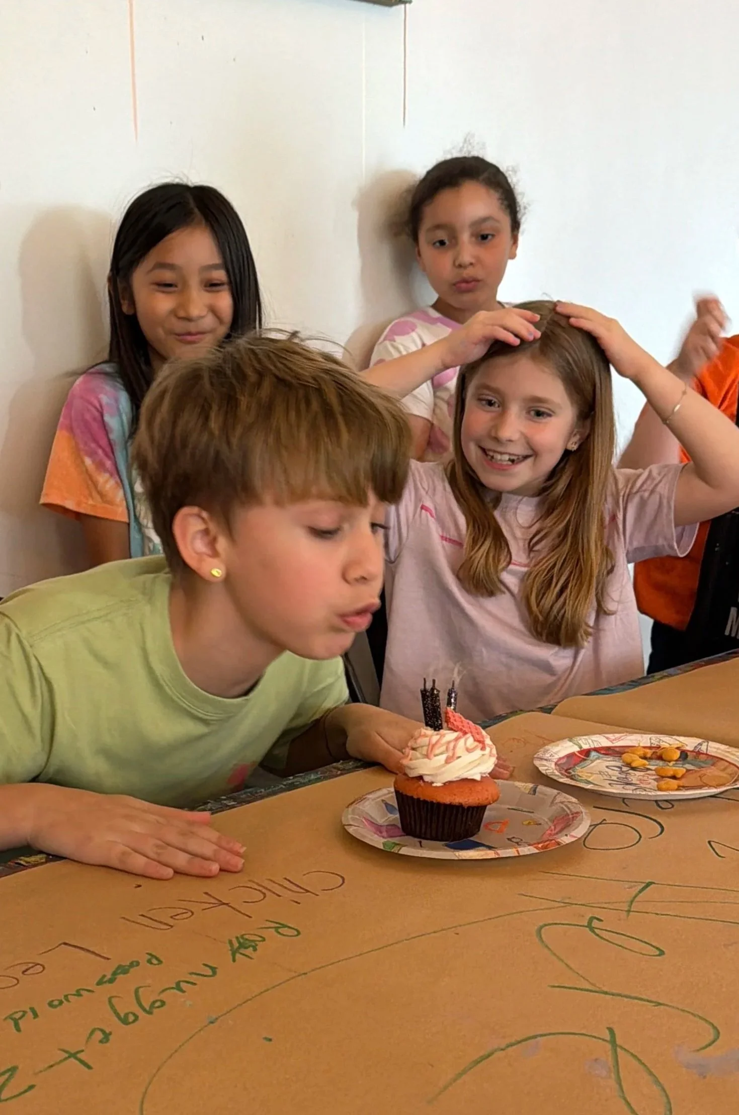 Children celebrating a birthday with a cupcake and small yellow snacks on the table.